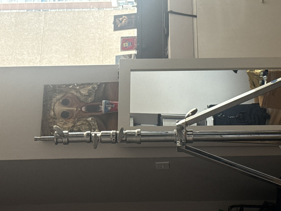 A low-angle shot of a metal stand in a room. A canvas painting featuring a figure with dark eyes and a tube of "ORAJET" is on a shelf. A white-framed mirror reflects parts of the room. On the wall, two posters appear above the painting. A window is visible in the background with a building exterior seen through it.