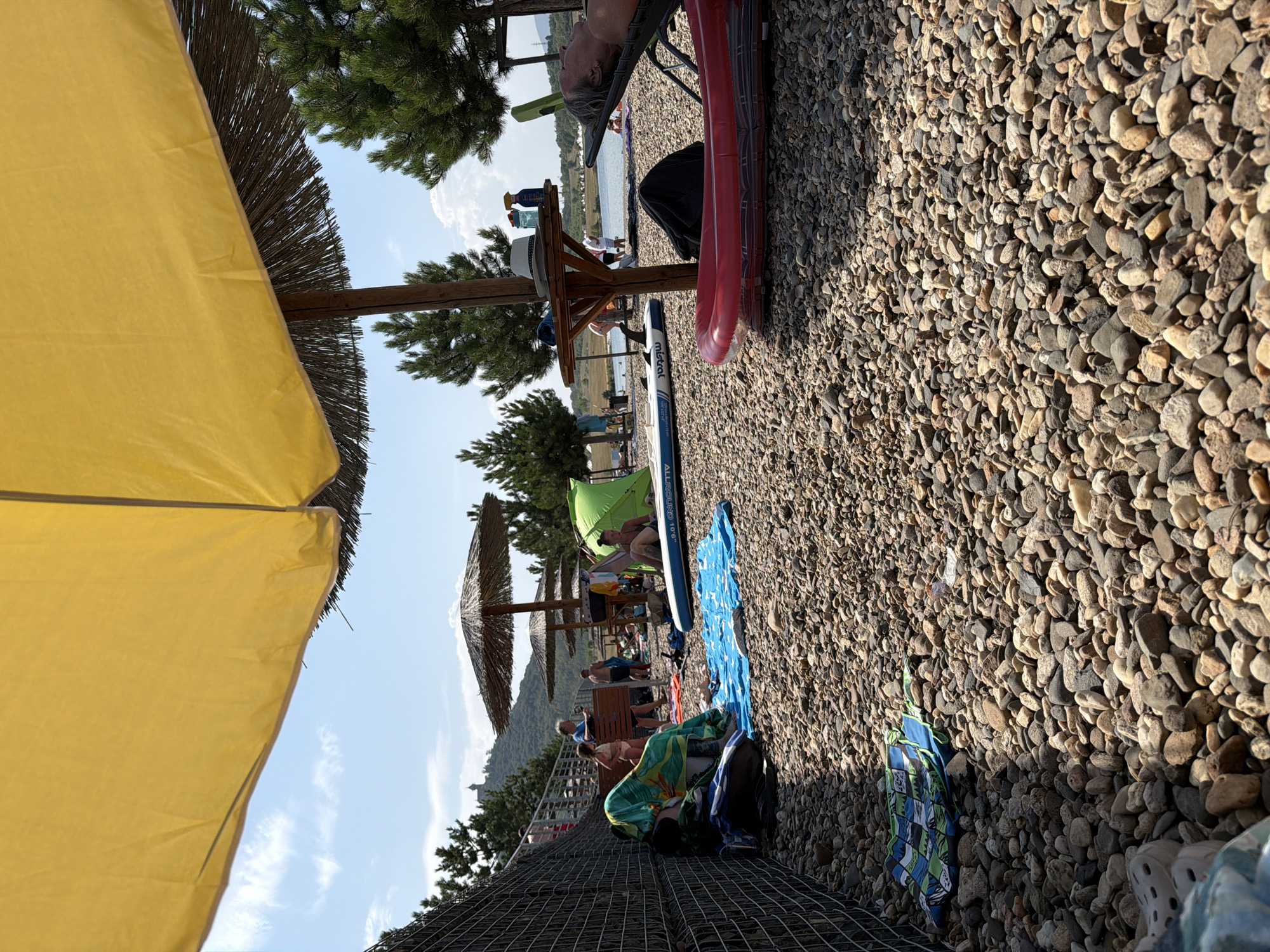 The image depicts a crowded beach scene on a sunny day, likely at the Most Reservoir in Most, Czechia. In the foreground, a large yellow umbrella partially obstructs the view of a pebble beach covered with various items. To the left, a black bag and a colorful towel are placed near a wire fence. Further back, people are seen relaxing under thatched umbrellas, some wading in the water, and others sunbathing on lounge chairs. A green tent is set up on the beach, and a paddleboard lies near it. In the background, lush green trees dot the landscape, and a body of water, presumably the reservoir, stretches towards the horizon. A building or structure is visible on a hill in the far distance. The overall atmosphere is one of a busy summer day at a recreational area.