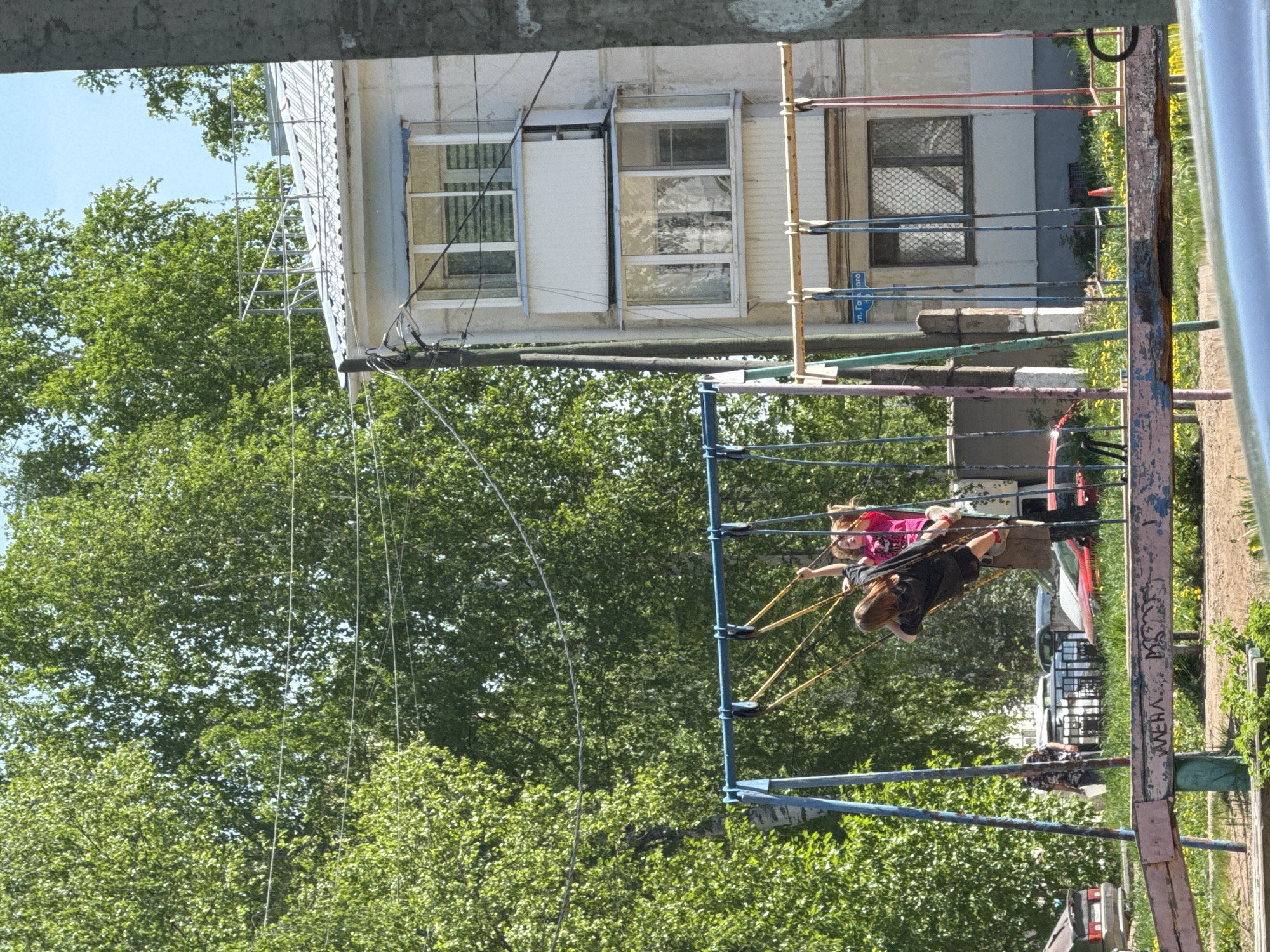 Observed from under a concrete overhang, the scene reveals a playground structure in Chaikovsky, Russia, located near a building with visible fire escape features. Two children are actively using the playground ride; one is wearing a pink shirt, while the other is dressed in black. In the background, cars are parked, and dense green trees provide a backdrop to the playground area. Graffiti marks one of the playground's support structures.