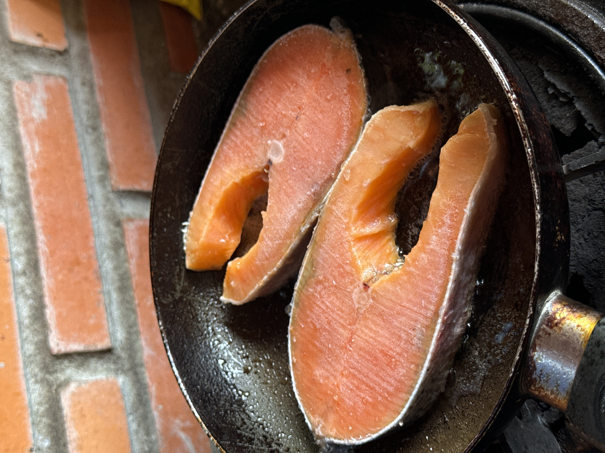 Two salmon steaks are being fried in a black frying pan. The pan sits on a black gas stove. A brick wall is visible on the left side of the image. The location is suggested to be Mũi Né, Vietnam.