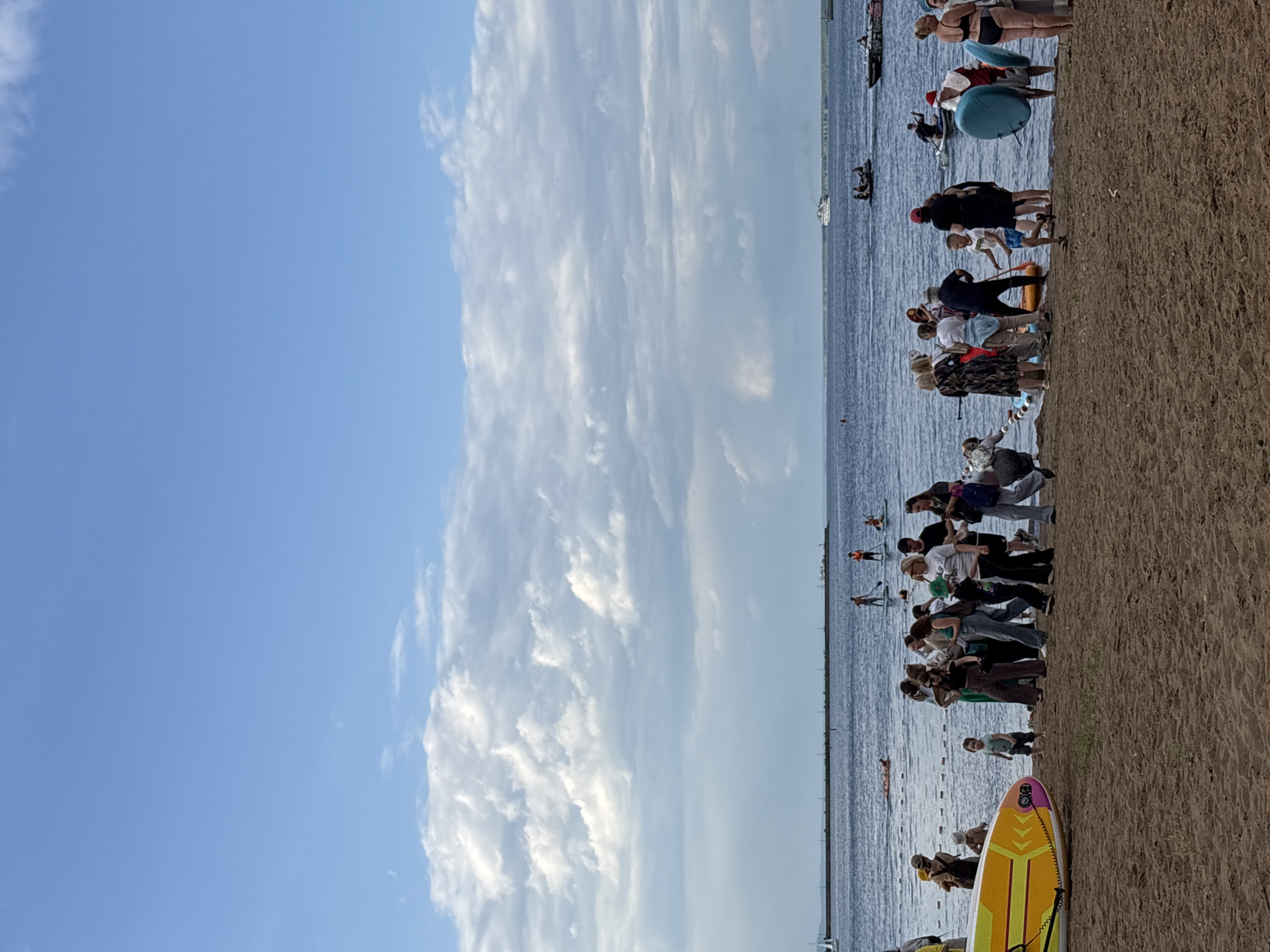 The image depicts a sunny day at a beach with a crowd of people gathered near the water. The scene appears to be a communal event or gathering, possibly a sporting activity or recreational day. There are many individuals, including adults and children, with some standing on the sandy shore and others in the water.
Several people are seen with paddleboards, suggesting a paddleboarding event or activity. Some individuals are standing on the boards in the water, while others are preparing to enter or exit the water with their boards. There are also what appear to be kayaks or canoes in the water, with people in them.
The sky is a bright blue with scattered white clouds, indicative of good weather and likely daytime, possibly late morning or afternoon. The water is calm, reflecting the sky. The beach is sandy, with some small waves lapping at the shore.
In the foreground, a colorful paddleboard is visible on the sand, with its fins and leash clearly discernible. The paddleboard has yellow, pink, and orange designs.
The people in the image are dressed in casual summer clothing, further supporting the idea of a warm, outdoor event. Their postures suggest a mix of participation and spectating. Some appear to be engaged in conversation or instruction.
In the background, a white boat or ship is visible in the distance on the water, along with a thin line of structures along the horizon. The overall atmosphere seems relaxed and festive, with a sense of community engagement. No text is clearly visible in the image.