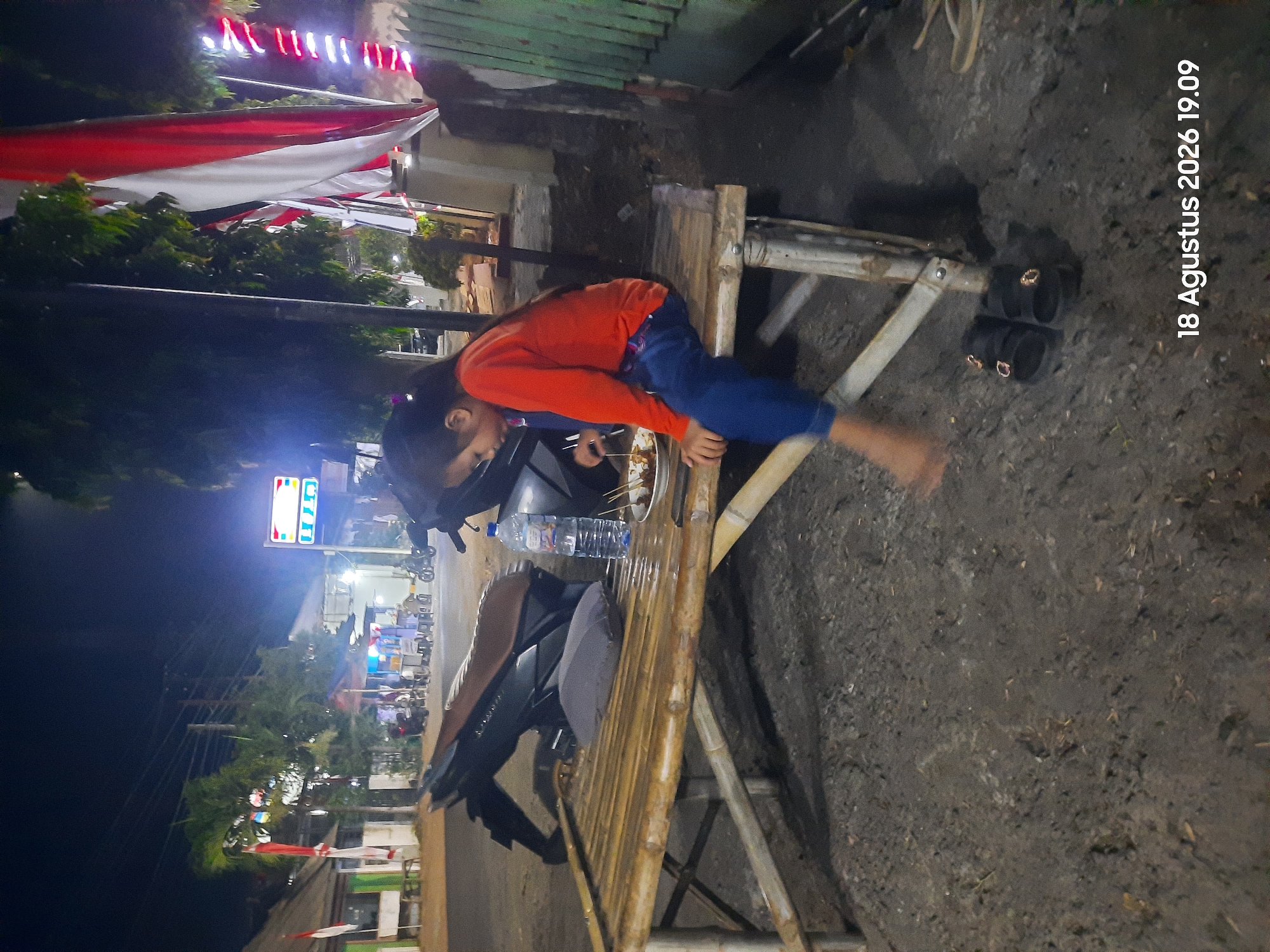A young girl in an orange long-sleeved shirt and blue pants sits on a bamboo bench at night in Sukokerto, Indonesia. She is looking down at a plate of food in front of her, which contains skewers and possibly meat. A bottle of water and a gray cushion are also on the bench. The girl's feet are bare, and she is wearing black sandals nearby. Behind her, a motorcycle is parked. In the background, there are buildings, trees, and Indonesian flags hanging. Streetlights and other illuminated signs are visible, suggesting a commercial or public area. The ground appears to be unpaved and dark. A timestamp in the bottom right corner reads "18 Agustus 2026 19.09."