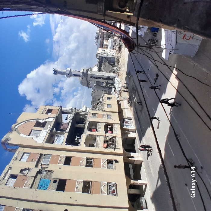The image presents a devastated urban street scene in Khan Yunis, Palestinian Territory, viewed from an elevated position. The sky is largely clear and blue with scattered white cumulus clouds. In the foreground, multiple dark electrical or communication cables span horizontally across the frame, partially obscuring the lower half of the image.
On the left, a multi-story building, appearing to be residential or mixed-use, displays significant structural damage. Several upper floors have large sections of their external walls missing, revealing interior spaces. A blue tarp is visible covering a window opening on the fourth floor from the bottom. Clothes are draped over a railing on a third-floor balcony. At street level, a light-colored sedan is parked on the sidewalk, and a group of three individuals—two adults and one child—walks along the sidewalk.
The paved street below features a faded white centerline. Approximately ten individuals, predominantly young males, are distributed across the street. Some appear to be engaged in activity with a ball.
In the background, a mosque with a tall, slender minaret stands amidst widespread rubble. The mosque's dome shows signs of damage, and extensive piles of concrete and building debris surround its base, indicating the destruction of adjacent structures. The minaret itself appears largely intact. The scene is characterized by widespread structural damage and debris, with ongoing civilian presence on the street.
