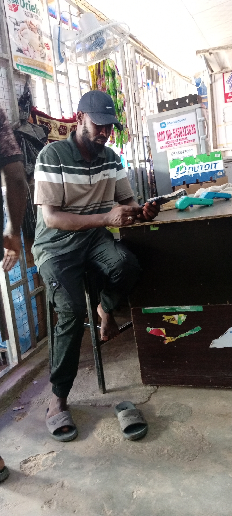 A man wearing a dark baseball cap and a striped polo shirt sits on a stool in what appears to be an outdoor market stall or small shop. He is looking down at a mobile phone he holds in his hands, his expression unreadable. He is wearing dark cargo pants and light grey slip-on sandals. To his right, on a wooden counter, are two electronic payment devices, one green and one teal. Behind him, a sign displays "Moniepoint ACCT NO: 5450323636 ACCOUNT NAME: BACHAKA SUPER MARKET". Another sign to the left advertises "Uriella FROZEN FOOD". Colorful streamers hang in the background, and a ceiling fan is visible above. The ground is concrete, showing some wear and tear. The scene suggests a transaction is either occurring or about to occur, or perhaps the man is simply using his phone during a lull in business. The lighting indicates it is daytime.