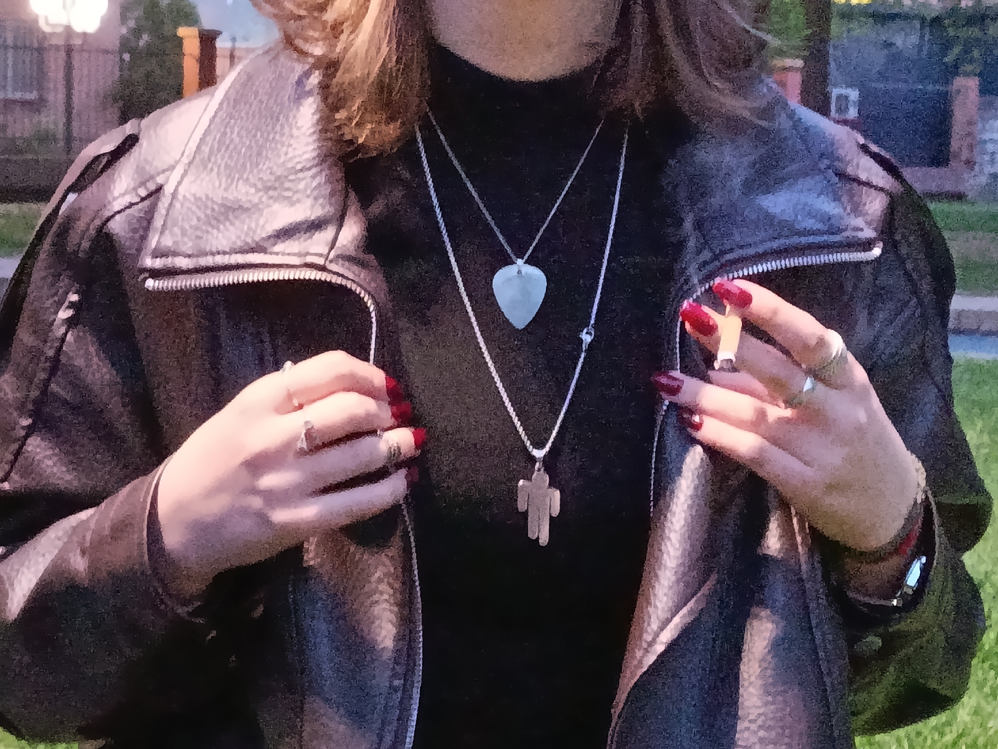 A young person stands outdoors, wearing a black turtleneck sweater and a black leather jacket. They are adorned with two necklaces: one with a heart-shaped pendant and another with a figure pendant. The person is holding a cigarette in their right hand, with visible rings on their fingers. Their nails are painted a dark red color. The background suggests an urban setting, possibly in Yerevan, Armenia, with buildings and greenery visible.