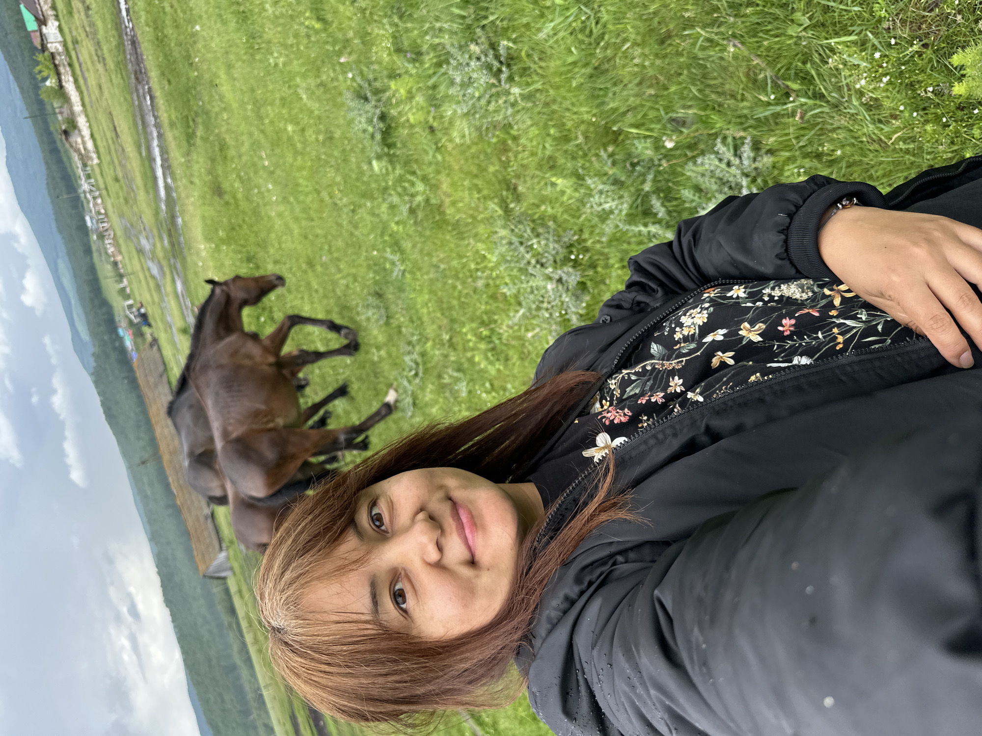 A woman with brown hair, wearing a black jacket over a floral shirt, takes a selfie in a grassy field. In the background, two horses graze in a muddy field, with a village and rolling hills in the distance. The sky is overcast. The scene is likely in Abzakovo, Russia, a popular resort town known for its natural beauty and horse riding opportunities.