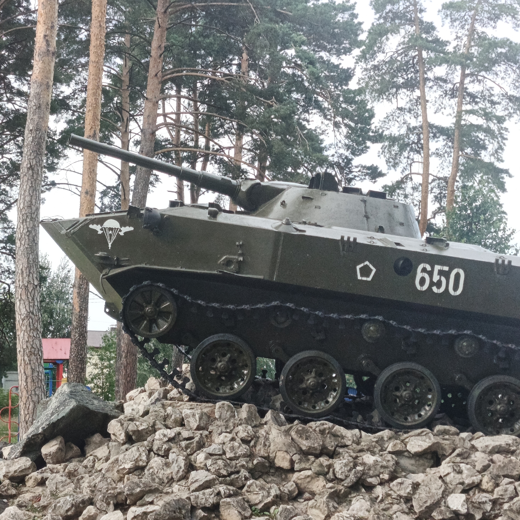 A military tracked vehicle, likely a Soviet-era BMP-1 or similar amphibious infantry fighting vehicle, is displayed on a mound of rocks outdoors, possibly as a monument. The vehicle is painted olive green and bears the number "650" and a pentagonal outline marking on its hull. A parachute insignia is visible on the front left side of the vehicle. The vehicle is situated amongst tall pine trees, suggesting a natural or park-like setting. In the background, a red-roofed building and playground equipment are partially visible, indicating a nearby residential or public area. The sky is overcast. The image composition is a medium shot focused on the vehicle's side profile.