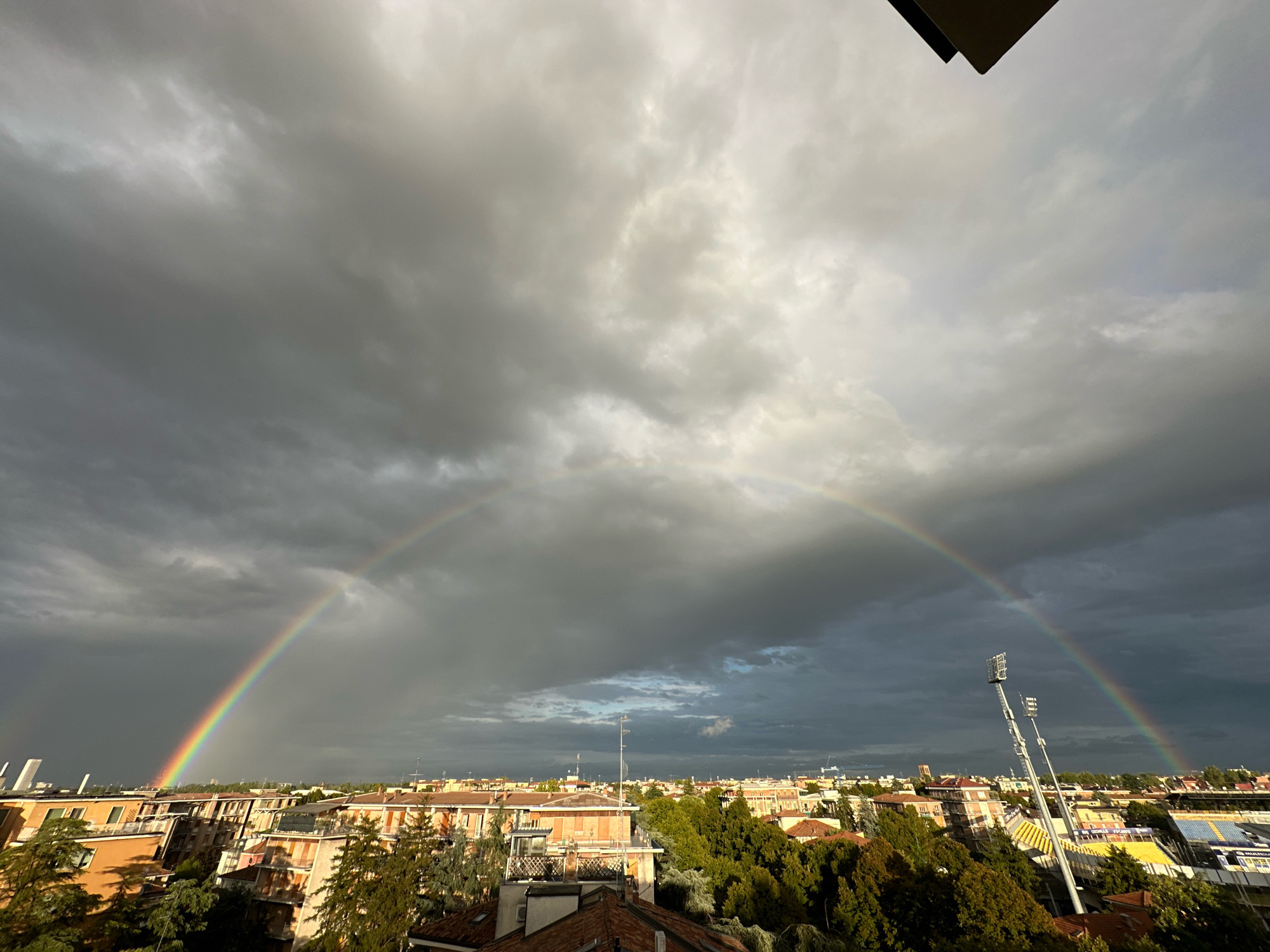 This image captures a vibrant double rainbow arcing across a cloudy sky over the city of Parma, Italy. The foreground shows a dense urban landscape with red-tiled roofs of residential buildings interspersed with lush green trees. The architecture suggests an Italian city, with the terracotta hues of the buildings being characteristic. In the mid-ground, a stadium with bright yellow and blue seating is visible, along with tall floodlights. The sky is dramatic, filled with heavy, dark gray storm clouds on the left, which transition to lighter, more diffused clouds on the right, allowing sunlight to break through and illuminate the rainbows. The rainbow on the left is more complete and brightly colored, while the secondary rainbow on the right is fainter. The scene appears to be taken from an elevated vantage point, possibly a balcony or rooftop. There are no people visible in the image.