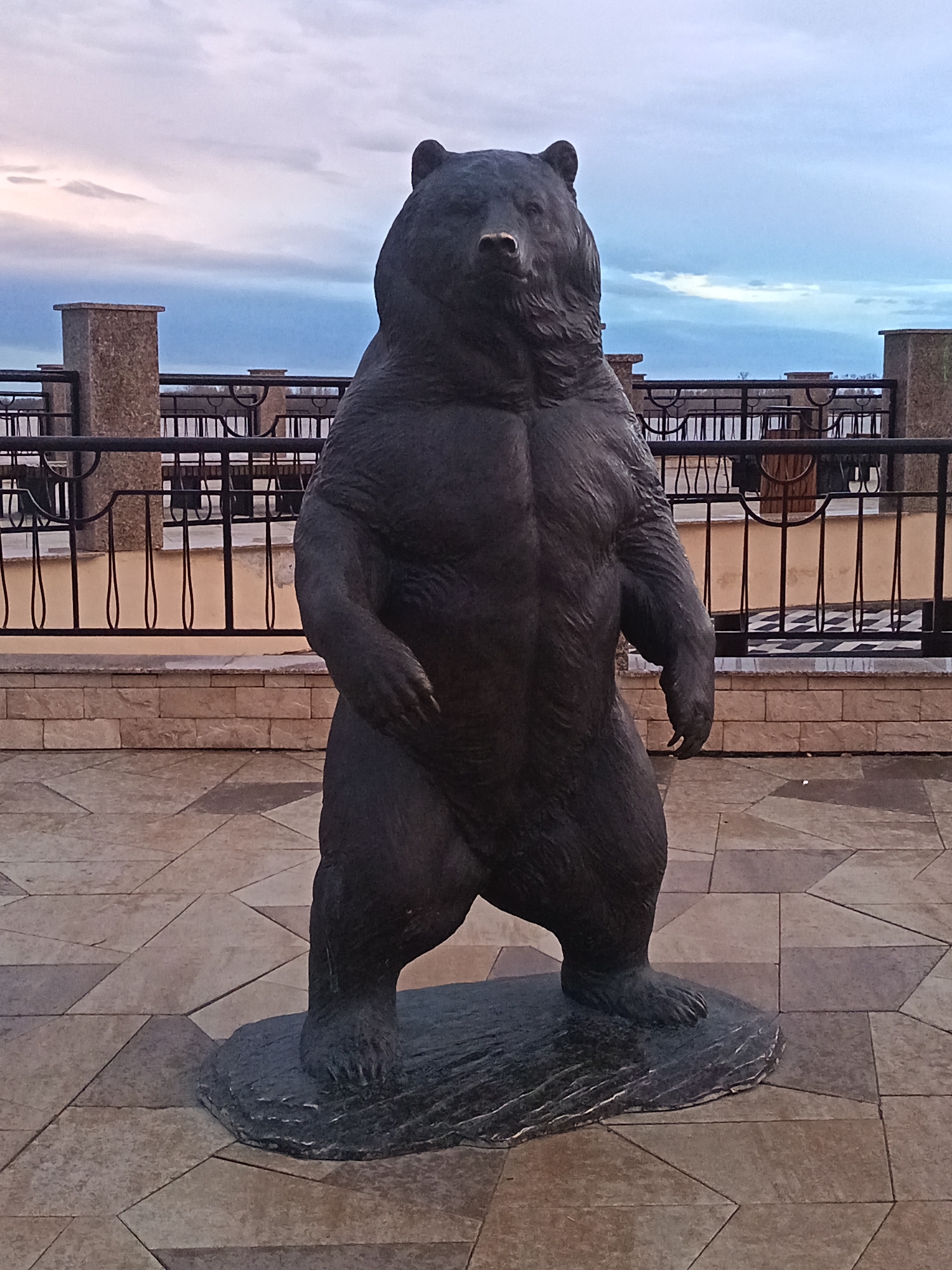 The image depicts a bronze statue of a standing bear. The bear has a muscular build and human-like posture. It stands on a small, textured base. The background features a pedestrian area with patterned pavement, a low brick wall, and a black metal railing. Beyond the railing, there are benches and a glimpse of a body of water under a cloudy sky. This statue is located on the embankment of the city of Nizhnevartovsk, Russia.