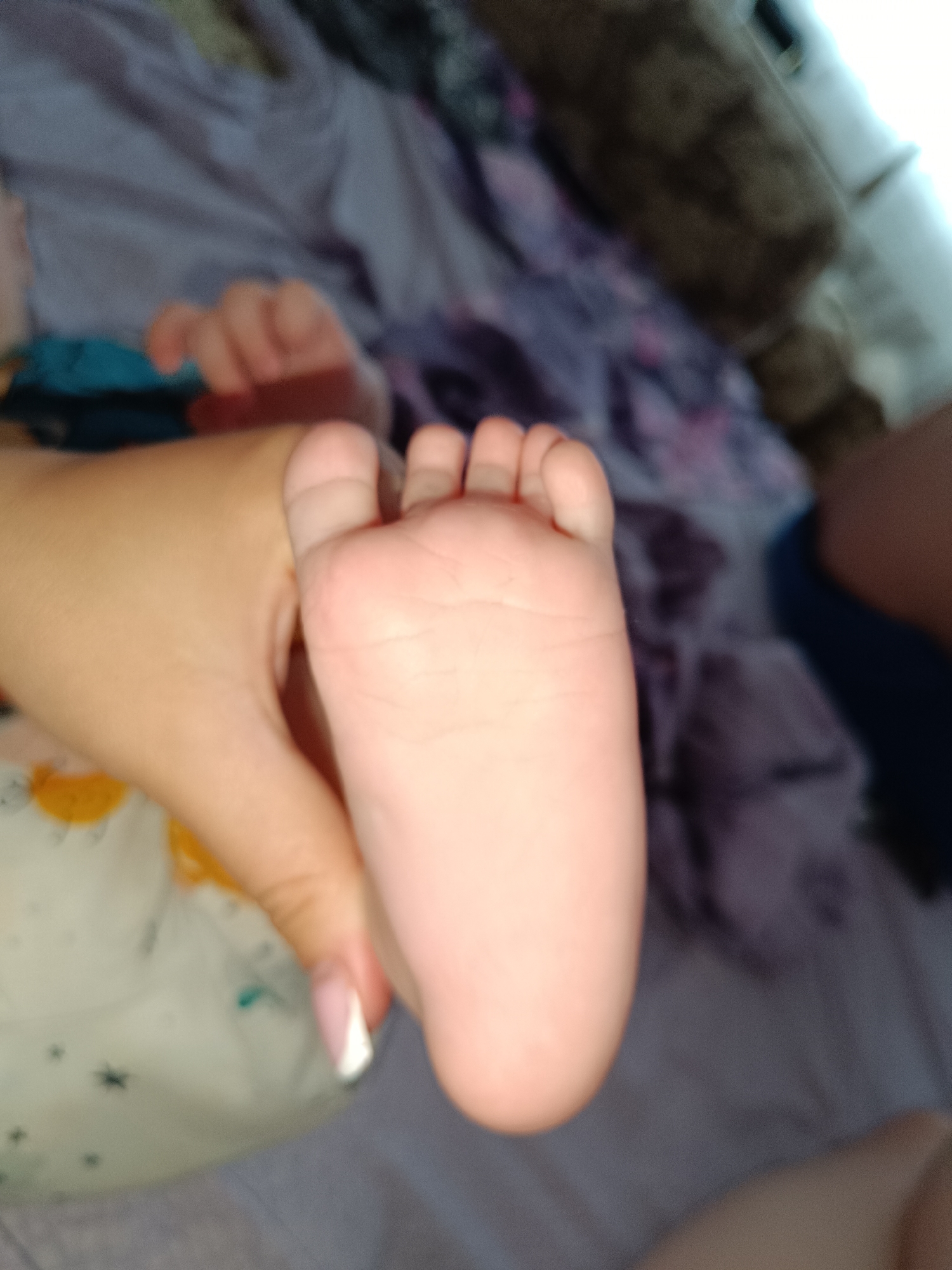 A baby's foot is being held in an adult's hand. The baby's foot is pink and chubby, with five toes visible. The adult's hand is fair-skinned and has a manicured nail with a white tip. The background is a blurred image of a bed with purple and white floral sheets. There is no evidence to suggest the location is Tobolsk, Russia.