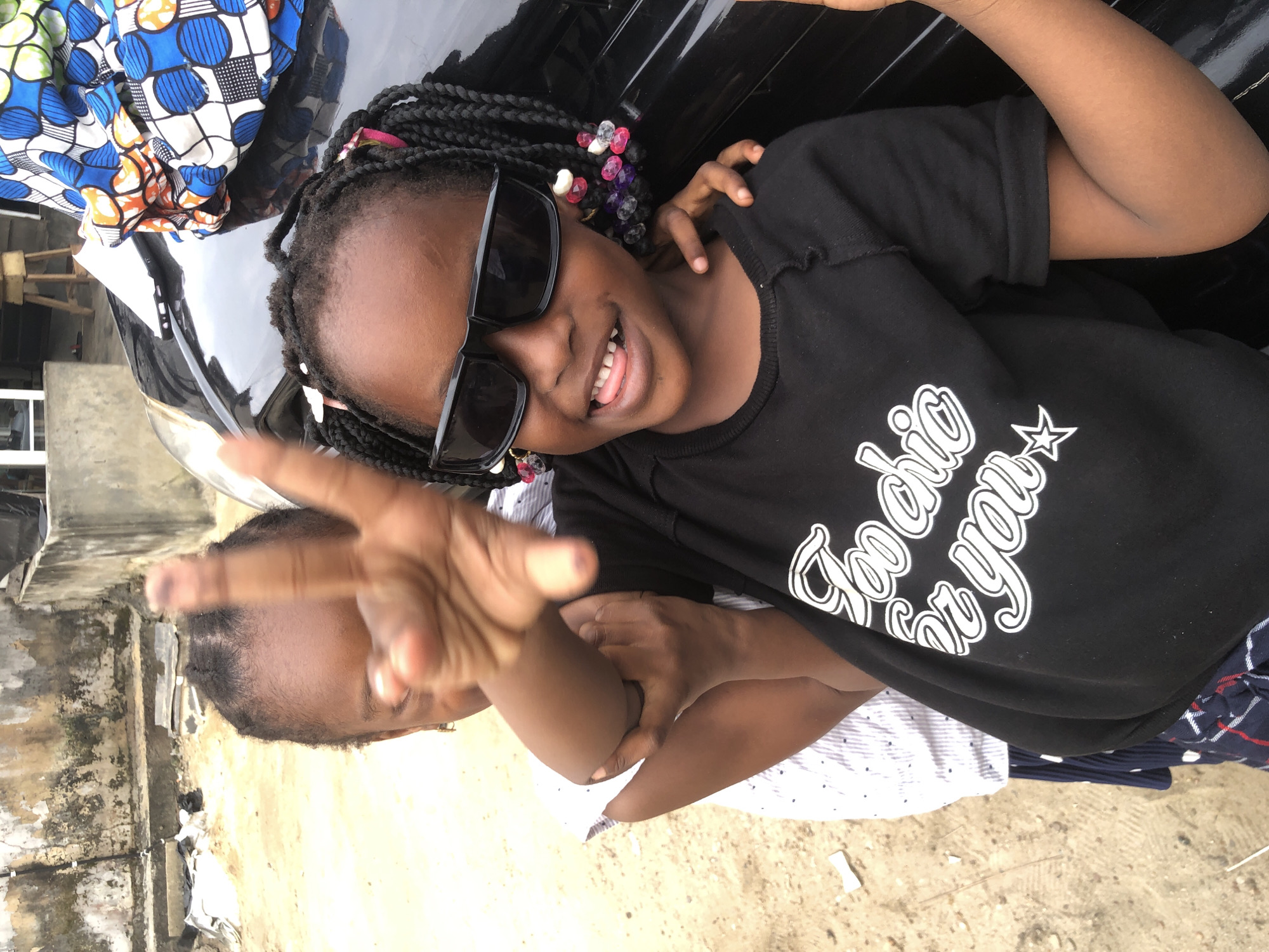 In the city of Ado, Nigeria, two young girls stand in front of a black vehicle. The girl in the foreground, wearing sunglasses and a black t-shirt with white text that reads "Too chic for you," playfully sticks out her tongue. Her braided hair is adorned with colorful beads. To her left, another girl, partially obscured, wears a white and blue striped shirt. The background shows a weathered wall and sandy ground.
