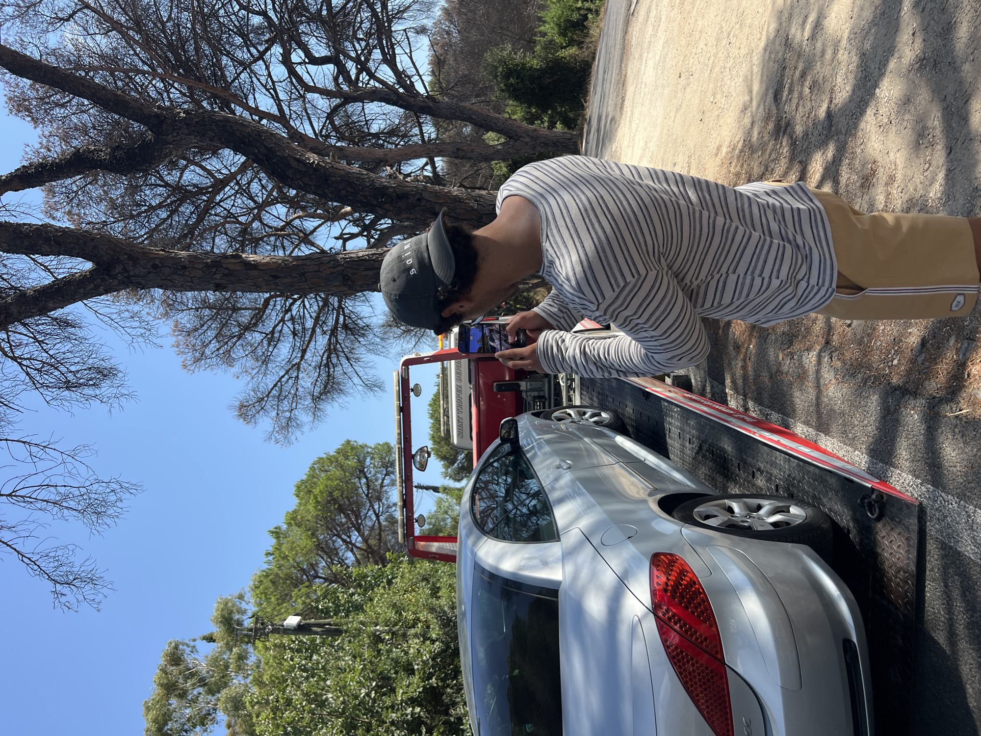 In Ramatuelle, France, a young man wearing a grey and white striped long-sleeved shirt, tan shorts, and a dark baseball cap is using his smartphone to take a photo of a silver convertible car being loaded onto a red tow truck. The car is positioned on the tow truck's platform. The scene is outdoors, with a clear blue sky and dry, brown tree branches overhead. The ground is dirt and gravel. Trees and foliage are visible in the background.