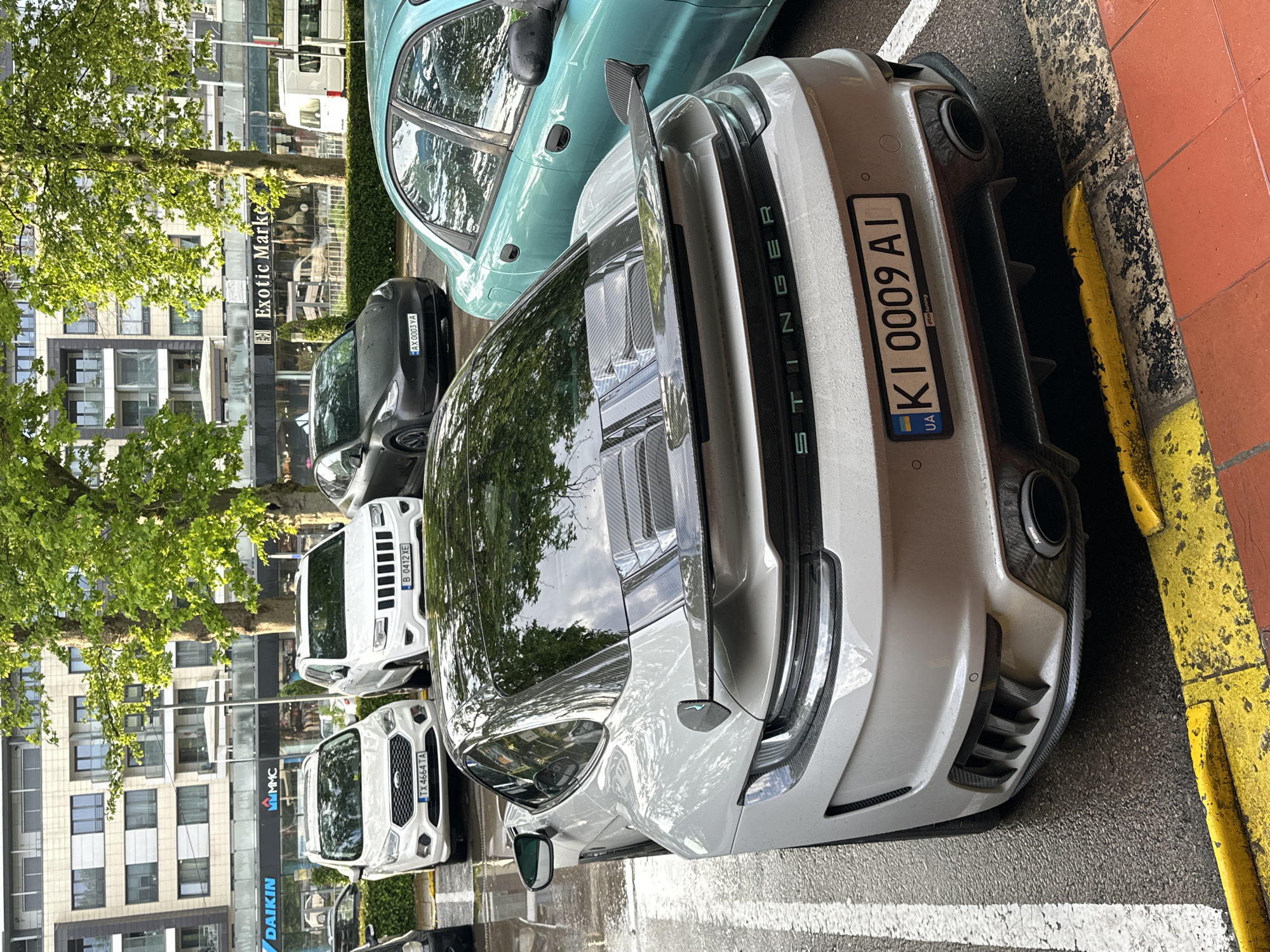 The image shows a street scene in Varna, Bulgaria, with several parked cars. The most prominent car is a silver modified Kia Stinger with a Ukrainian license plate reading "KI 0009 AI," parked close to a curb painted with yellow and black stripes. To the left of the Stinger, other vehicles are visible, including a white Ford Transit with the license plate "TX 4664 IA" and a white SUV, parked in a row. In the background, buildings with businesses are visible, including a Daikin air conditioning office and a sign reading "Exotic Marke." A black car with the license plate "AX 0003 YA" is parked next to a turquoise car. Trees with green foliage are situated along the street, casting reflections on the vehicles. The street appears wet, suggesting recent rain.