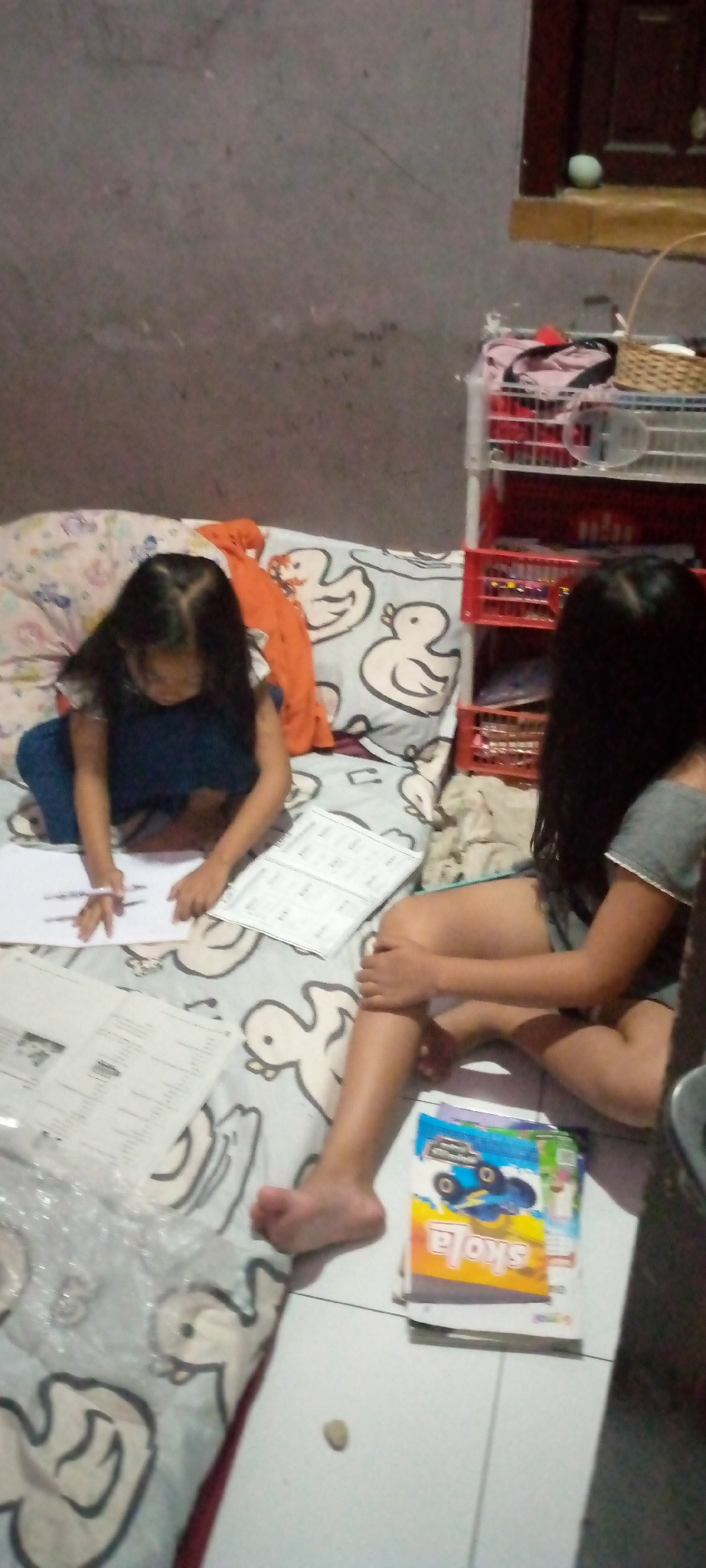 In a domestic indoor setting, likely a bedroom, in Sumberwono, Indonesia, two young girls are engaged in an activity. One girl, seated on a bed with a pillow featuring a duck motif, is wearing a dark blue dress and is focused on writing with a pen on a white sheet of paper. Adjacent to her is another open booklet. Beside her, on the bed, is an orange garment. To the right, another girl, wearing a grey t-shirt and dark shorts, is seated on the floor, her legs extended. She is holding her knee with her left hand. In front of her, a stack of books, one with a blue monster truck graphic and the word "SKOLA," rests on the floor. The environment appears to be dimly lit, with a textured, grey wall in the background. A red shelf unit with multiple compartments is visible to the right, containing various items. A small, light-colored stone is on the floor in the foreground.