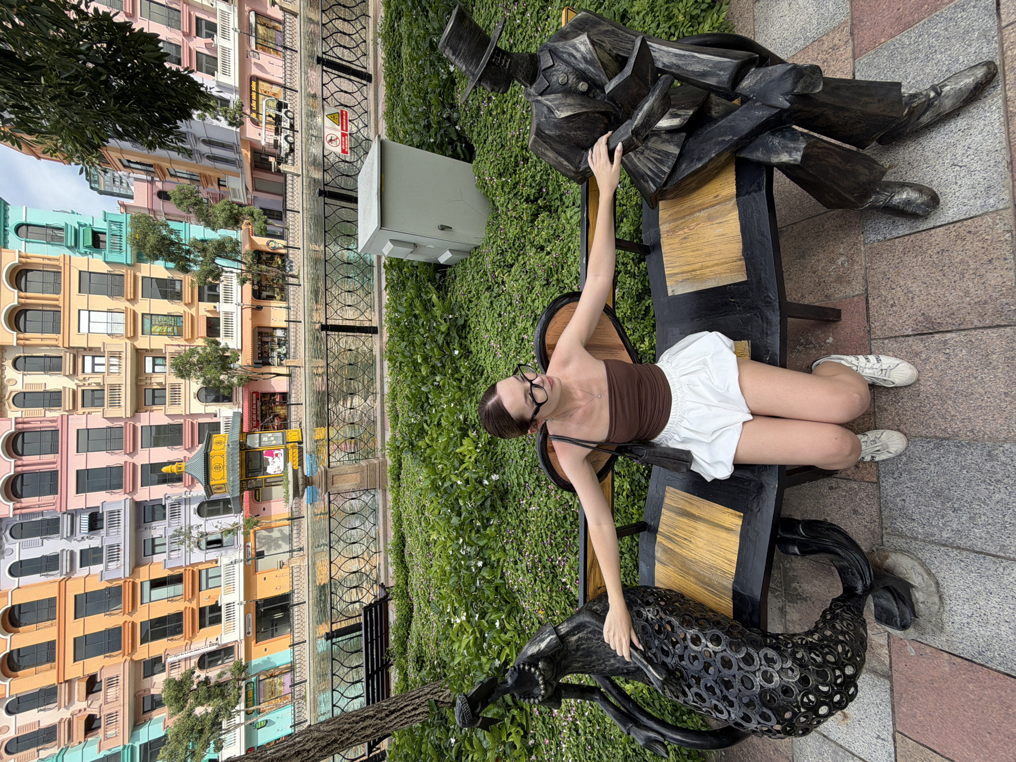 A young woman with brown hair, wearing glasses, a brown tube top, and a white skirt, sits on a black bench in a park. She is positioned between two black metal sculptures: one on her left depicting a woman with elaborate detailing on her dress, and another on her right of a man in a top hat and suit, holding what appears to be a book. Her arms rest on the sculptures. The bench has a wooden seat. Behind the bench is a dense green bush with small pink flowers. In the background, a body of water is visible, with a black metal fence separating it from the park. Beyond the water, a row of colorful buildings with arched windows and balconies line the waterfront. A utility box is situated in the bushes to the right of the bench. The paving stones in the foreground are a mix of red and grey. The overall scene suggests a vibrant, possibly resort-like, urban environment in Nghĩa Trụ Commune, Vietnam.