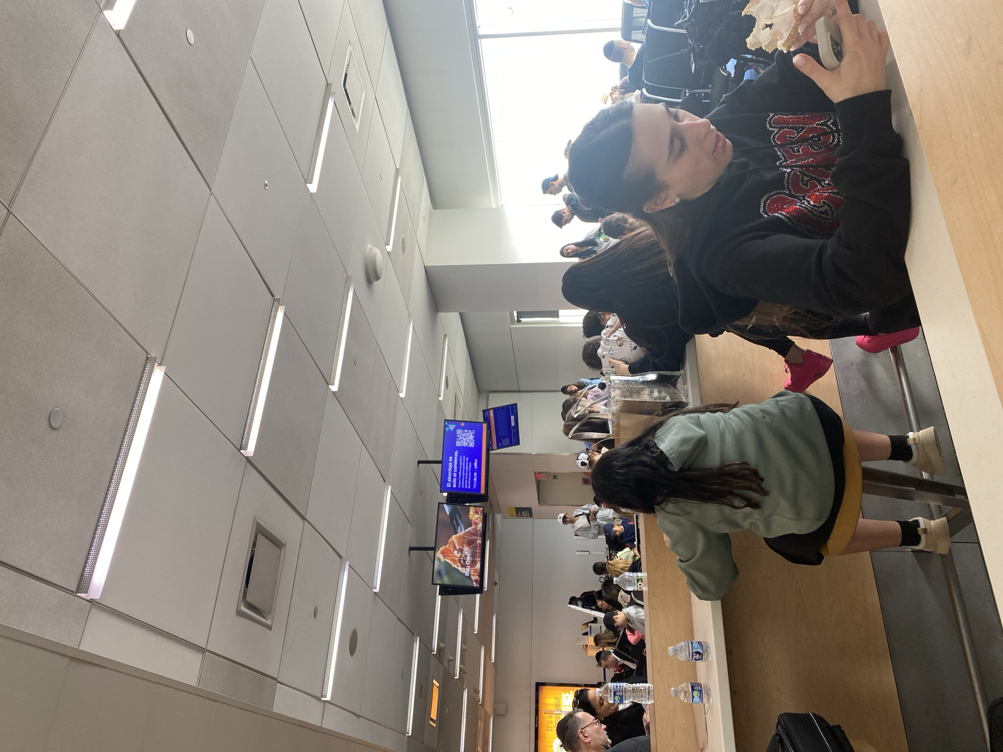 The image shows an indoor scene, seemingly a waiting area with multiple people seated at tables. The room has a modern design, characterized by a high ceiling with linear lighting fixtures. There are several digital screens mounted on the wall, displaying advertisements and information. Individuals are engaged in various activities, such as eating, talking, and using electronic devices. Water bottles are visible on the tables. The location is likely an airport terminal, possibly in New York, United States, due to the presence of the airline advertisements and the general layout resembling airport seating areas.