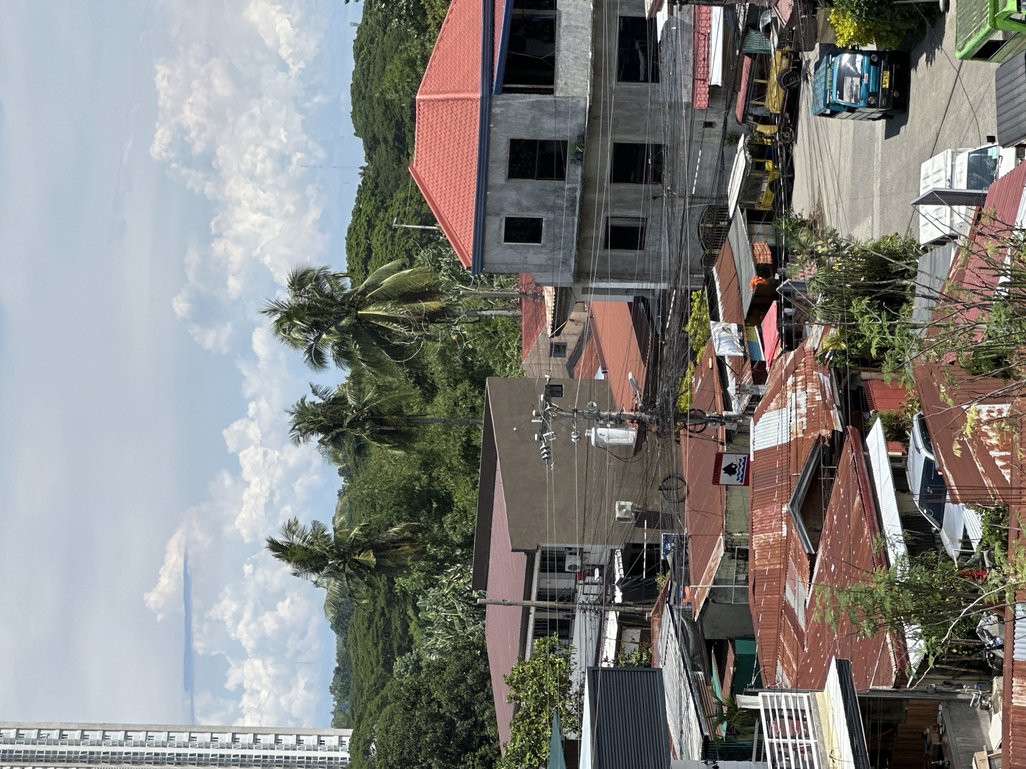 The scene is a high-angle view in Davao City, Philippines, showing a mix of residential and commercial buildings. A partially constructed gray concrete building with a red tile roof stands prominently. Below and adjacent to it are older structures with corrugated metal roofs, many exhibiting rust and weathering. A dense network of power lines crisscrosses the view, obscuring some of the buildings. A street is visible, with a jeepney, a white truck, and a white car parked along the roadside. The sky is partly cloudy, and lush green vegetation and palm trees are visible in the background, suggesting a tropical environment. A tall, multi-story apartment building is visible in the lower-left corner of the image.