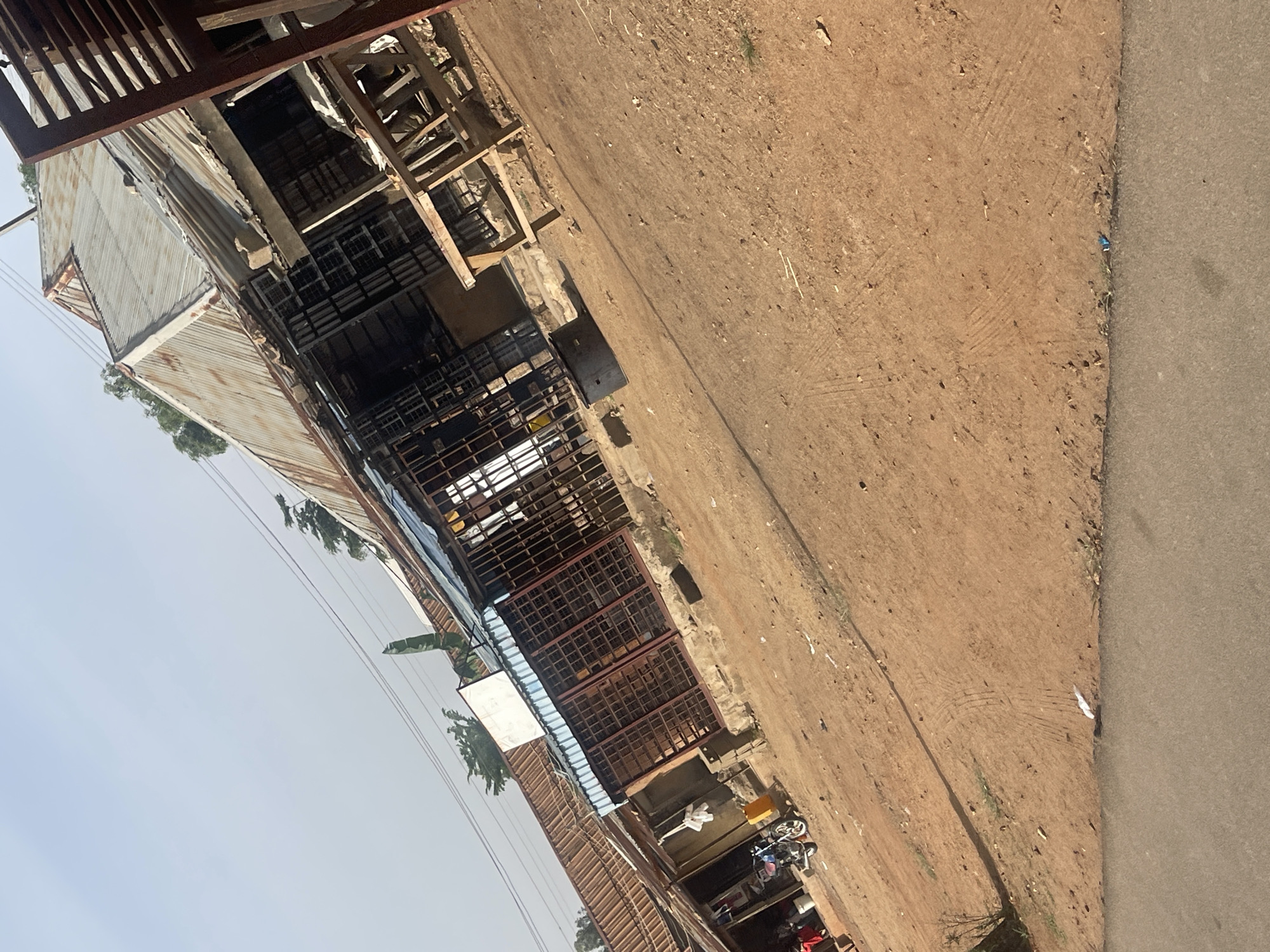 This image captures a daytime scene in Jos, Nigeria, featuring a street alongside functional buildings. The view is oriented with the ground on the right and structures on the left, under a clear sky.
On the left side of the frame, several buildings with weathered corrugated iron roofs are visible. Beneath these roofs, the structures feature multiple rectangular metal grates, likely serving as windows or security barriers for workshops or market stalls. Some wooden framework suggests ongoing construction or repair. A small, white, rectangular sign is also positioned near the center of these structures.
The right side of the image is dominated by a wide, reddish-brown dirt path that shows signs of use, including tire tracks and scattered small debris like stones and sticks. This unpaved area borders a narrower, darker gray paved road that runs parallel to the path on the far right.
Near the base of the buildings, on the dirt path, various objects are present. These include a white item that resembles a plastic chair or bucket, yellow containers, and what appears to be parts of a bicycle or motorbike, along with other indistinct items.
The sky in the upper left is clear and light, indicating bright daytime conditions. No people are visible, and no specific activity or event is clearly taking place, presenting a still snapshot of the environment. There is no discernible text visible in the image. The overall impression is that of a bustling, utilitarian street or market area.