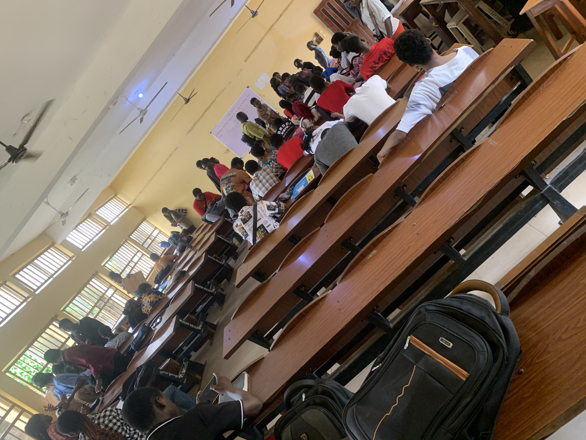 The image depicts a classroom scene with a group of students seated at wooden desks arranged in rows. The room has large windows with blinds on the left side, letting in natural light. Several ceiling fans are visible. The students are mostly young adults, dressed casually. Some are looking forward, possibly at a teacher or lecture, while others are engaged in conversation or using their phones. Backpacks and other personal belongings are placed on the desks or on the floor. The overall atmosphere suggests a lecture or class in session. The location is specified as Unknown, Unknown.