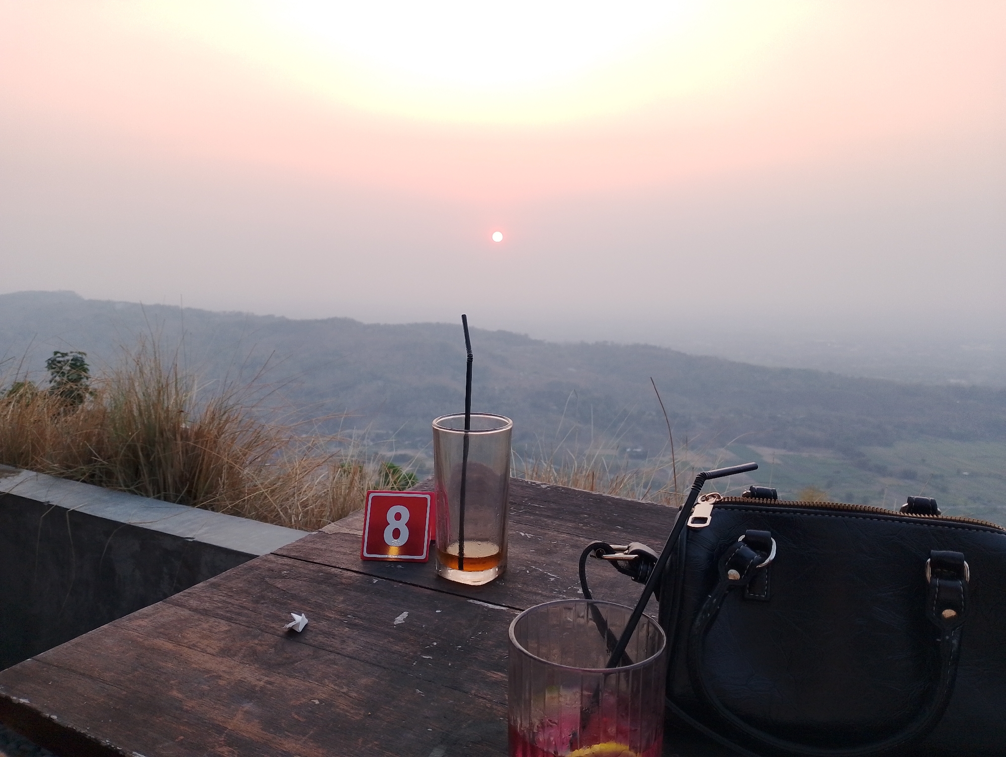 The image depicts a serene sunset scene at a mountaintop cafe overlooking Srimulyo, Indonesia. A wooden table is set with two glasses, one with a straw and a small amount of amber liquid, and another with a pinkish drink and a slice of fruit. A table number sign, displaying the number "8" in red, is placed next to the first glass. A black handbag rests on the table to the right, with a straw peeking out from the glass beside it. In the background, the sun is a small, bright orb in a hazy sky, casting a soft glow over rolling hills and distant landscapes. Tall grasses frame the left side of the scene, suggesting an outdoor setting. The overall atmosphere is tranquil and suggestive of a relaxing evening at a scenic viewpoint.