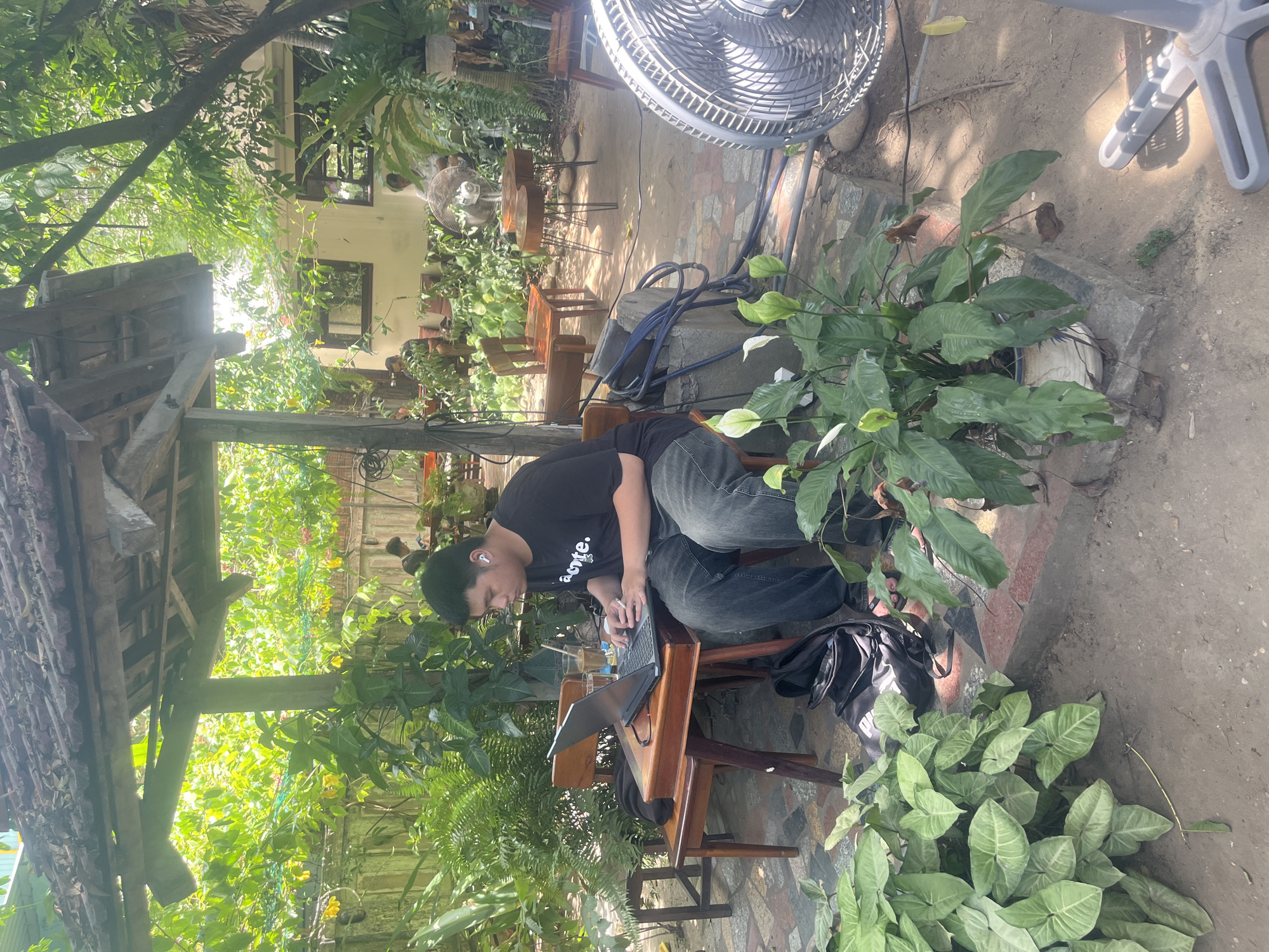 A young man is working on a laptop in an outdoor cafe in Da Nang, Vietnam. He is sitting at a small wooden table with a drink next to him. He is wearing a black t-shirt and jeans and has earbuds in. The cafe is filled with plants and has a rustic, open-air feel. There is a fan to the right of the image, and other patrons are visible in the background. The man appears focused on his work, with the dappled sunlight filtering through the trees overhead.