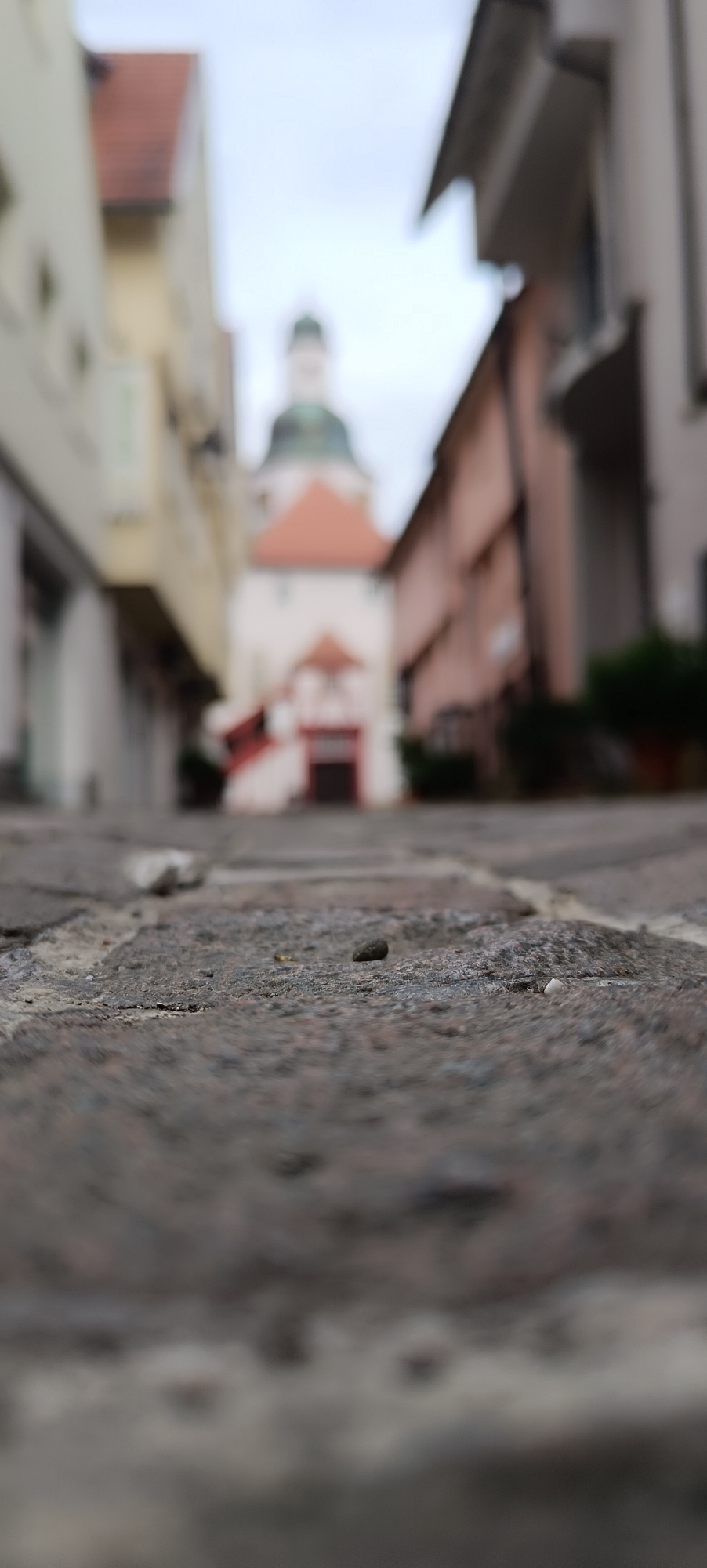 The image captures a low-angle perspective of a cobblestone street in what appears to be an old European town. In the foreground, the rough texture and irregular shapes of the cobblestones are sharply in focus, with a few small pebbles scattered on the surface.
The background is blurred, creating a shallow depth of field that draws attention to the street itself. The blurred elements suggest buildings lining a narrow street, with a church or steeple visible in the distance. The architecture includes elements like pitched roofs and facades that are typical of historic European towns. One building features a prominent orange-tiled roof, and another a rounded green dome, suggesting a church or religious building at the end of the street. The overall impression is of an overcast day, with soft, diffused light. There is no visible text in the image. The scene evokes a sense of quiet and perhaps a bit of solemnity, characteristic of a cloudy day in a historic urban setting. The location is identified as Winnenden, Germany.