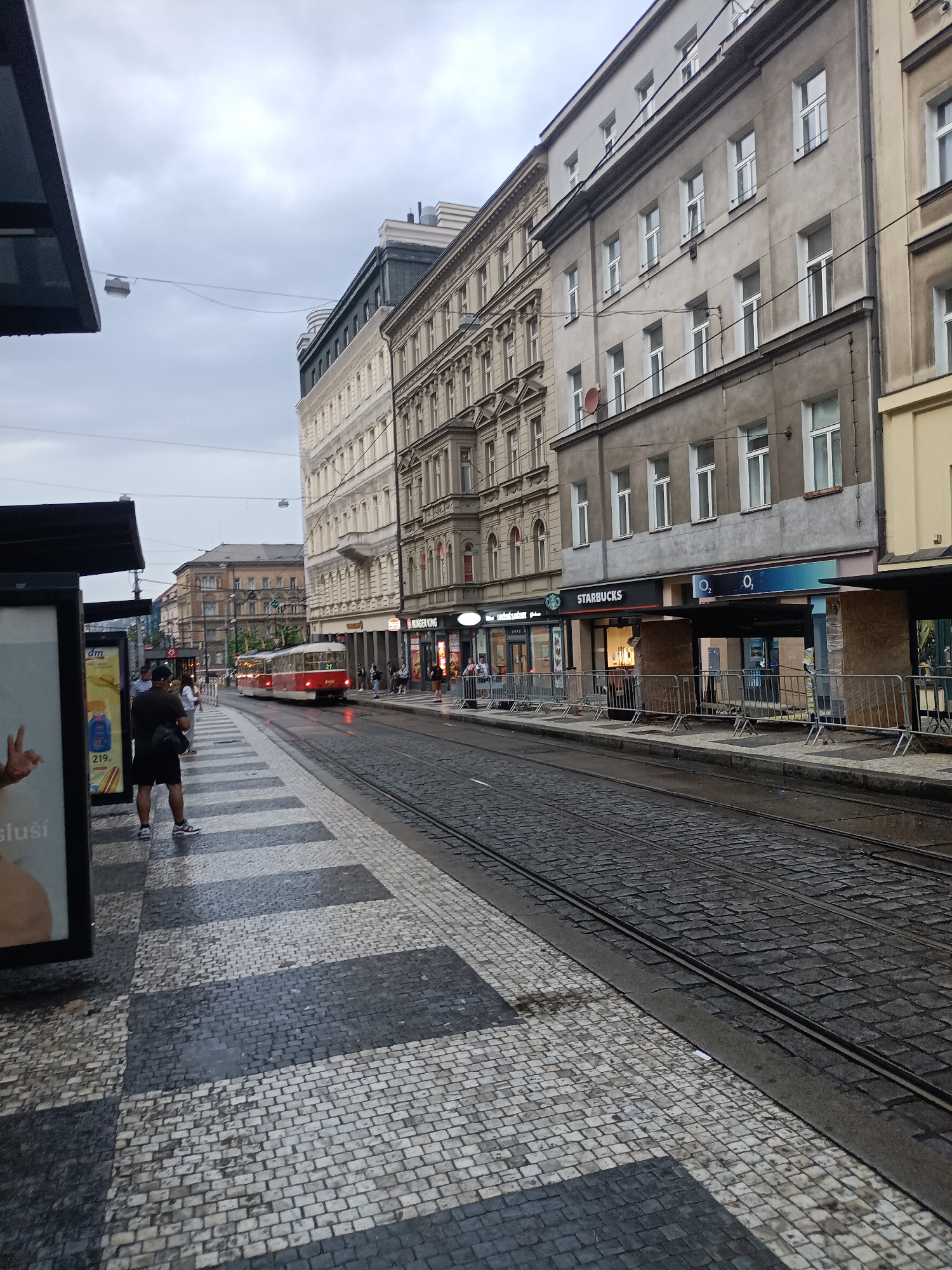 The image captures a view of a tram stop and street in Prague, Czechia, likely on a cloudy or overcast day. In the foreground, a man stands at the tram stop, facing away from the camera, wearing a dark t-shirt and black shorts. The tram stop features a patterned tiled platform with alternating dark and light grey squares. To the left, a black advertising display is visible. The street is paved with cobblestones, and tram tracks run along the center. A red and white tram is seen approaching in the distance. Along the right side of the street, buildings line the sidewalk, with storefronts visible including Starbucks and O2. Several people are walking on the sidewalk. The sky is overcast with a lamp post hanging from wires visible in the upper left quadrant.