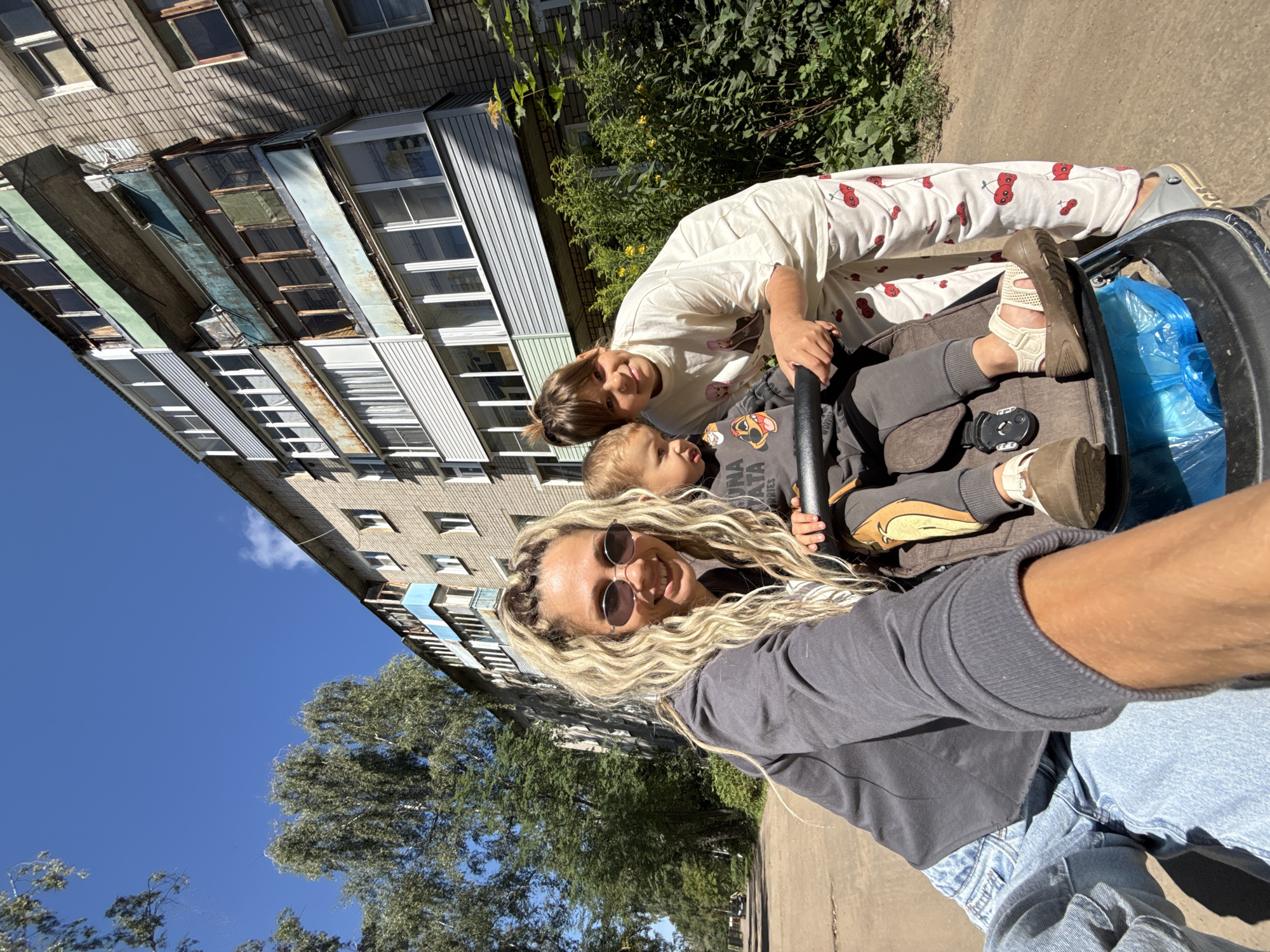 In Rusanovo, Russia, on a sunny day, a woman with blonde, braided hair and sunglasses takes a selfie while pushing a stroller. Next to her, a girl in a white t-shirt and cherry-patterned pants leans in. Inside the stroller, a young child sits, wearing a grey sweatshirt with cartoon characters. The background features a weathered, multi-story apartment building and lush green trees lining a dirt path. The overall scene captures a casual moment outdoors, with the urban apartment block providing a backdrop to the family's outing.