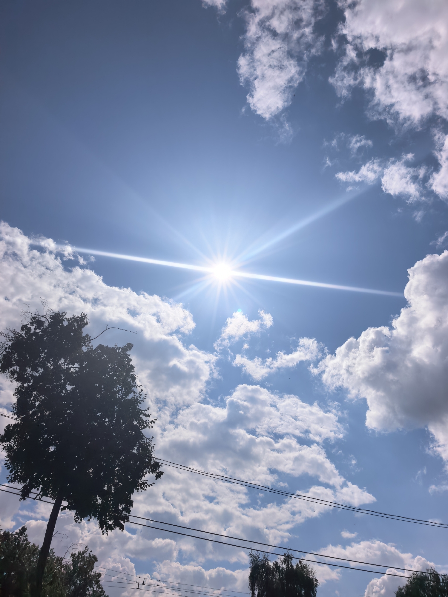 This image depicts a bright, sunny sky with scattered white clouds. The sun is a dominant feature, positioned in the upper center of the frame, emitting strong rays of light and causing a lens flare. A silhouetted tree with dark foliage stands on the left side of the image, its branches reaching towards the sky. Power lines run horizontally across the middle and lower portions of the frame. In the background, the tops of other trees are visible, suggesting a semi-rural or suburban setting. The sky is a vibrant blue, indicating a clear day. The presence of the sun high in the sky suggests it is likely daytime, possibly late morning or early afternoon. The overall mood is one of brightness and clarity, with a slight sense of vastness due to the expansive sky. The location is identified as Sterlitamak, Russia, which provides geographical context for the scene. There is no visible text in the image.
