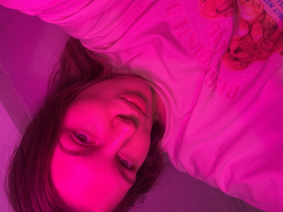 A person with fair skin lies on a gray couch, illuminated by a strong pink light. They have light brown hair, and their face is turned toward the camera. They wear a light-colored shirt, partially obscured by the pink light, with a teddy bear visible in the upper right corner. The shirt has text, "DISAPPOINTED CLUB" on it. The subject's eyes are open, and the pink light dominates the color scheme of the image.
