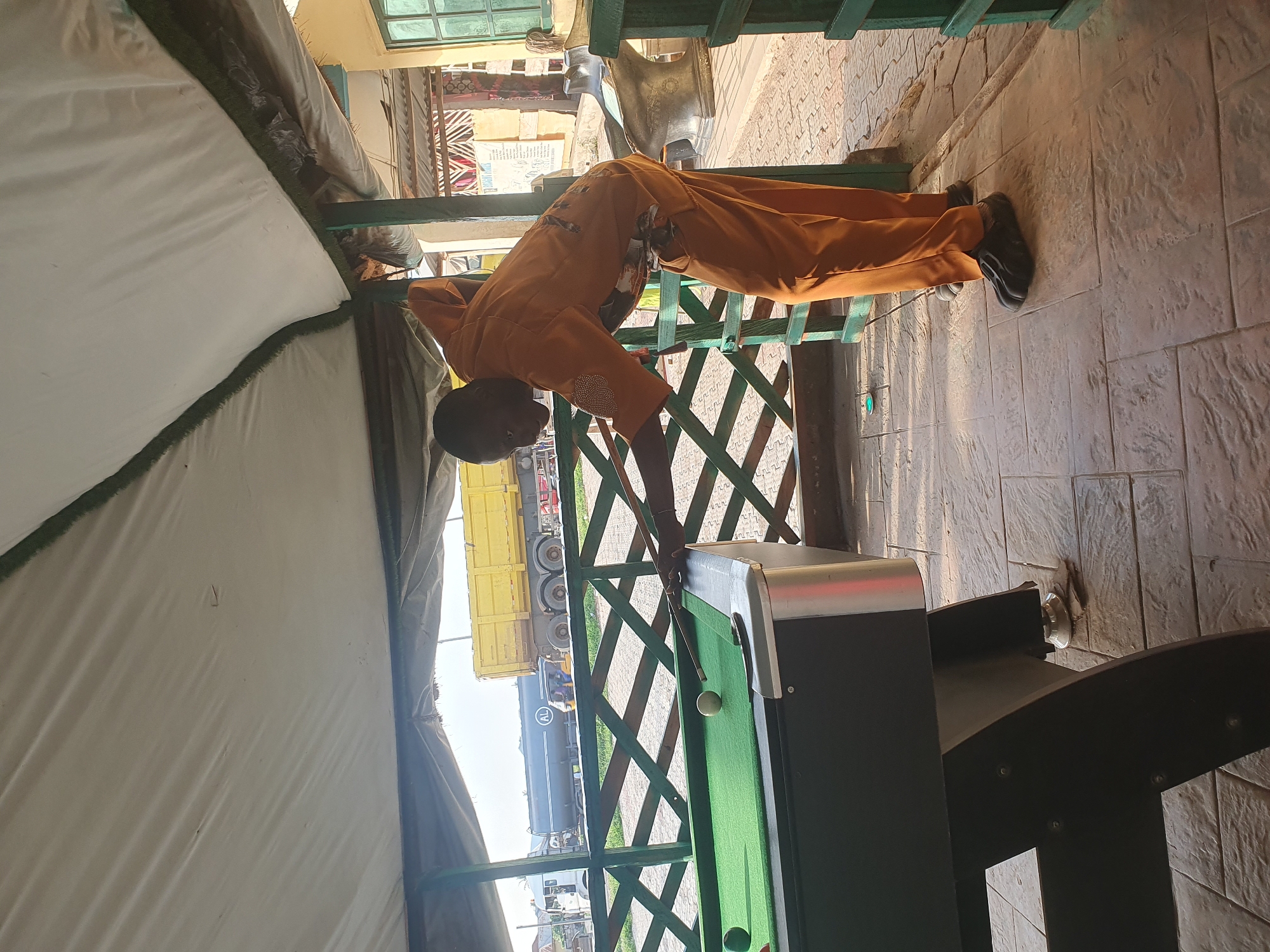 In Lokoja, Nigeria, a man in an orange jumpsuit plays pool under a white tarp canopy. The pool table, covered in green felt, has several balls on it. The man is bent over, aiming the cue stick at a ball. In the background, a yellow truck and a large tanker truck are visible on a road, along with some buildings and a green wooden fence. The ground is paved with textured tiles.