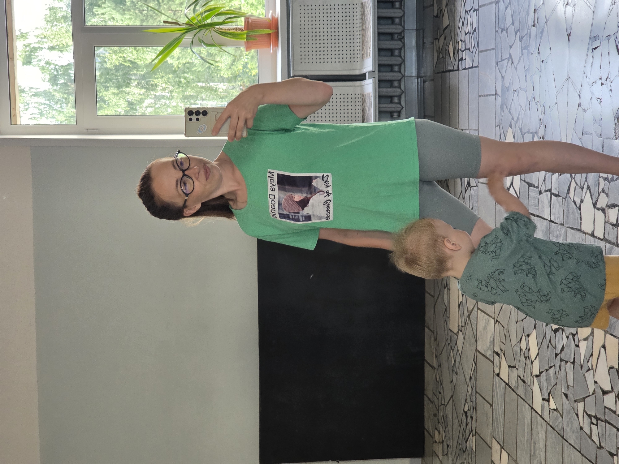 The image shows a woman and a young child in an indoor setting, possibly a room with a large window. The woman is wearing a green t-shirt with a picture on it and has glasses on. She is holding a smartphone, taking a selfie. The child is wearing a green t-shirt with dinosaur prints and yellow shorts. The background features a window with trees visible outside, suggesting daylight. The floor is tiled with a mosaic pattern. A radiator and some shelves are also visible. The overall mood appears casual and domestic. The text on the woman's t-shirt is partially visible and appears to be in a foreign language.