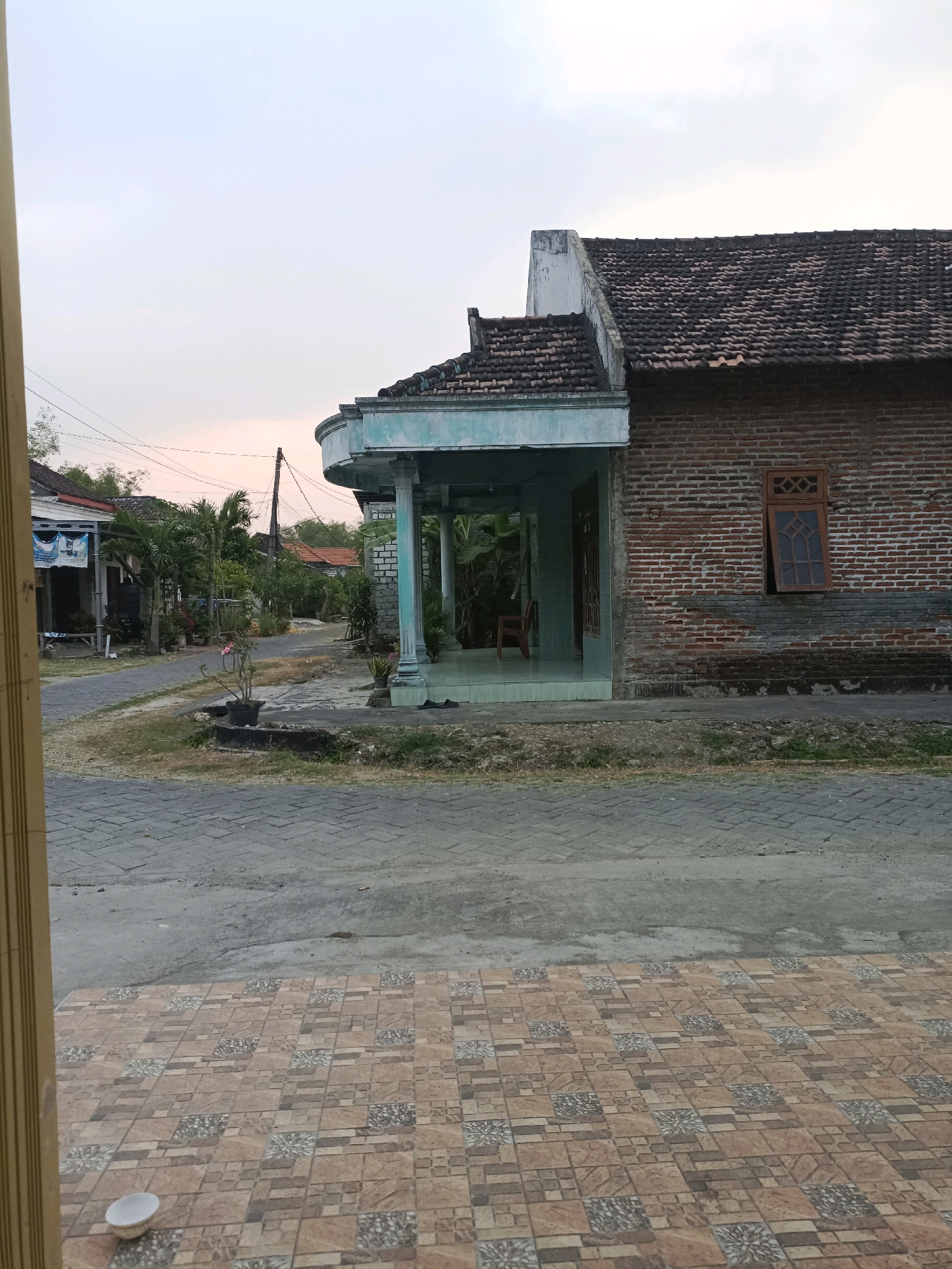 The image shows a street scene in Rancang Kencono, Indonesia. The foreground features a tiled patio with a white bowl. Beyond the patio is a paved road and a sidewalk. A house with a brick facade and a dark tiled roof is visible on the right side of the image. The house has a wooden window with a diamond-shaped pane. A covered porch with pillars extends from the house. To the left of the house, a street lined with houses and vegetation stretches into the distance. Power lines are visible across the sky. The sky is overcast with a soft, diffused light, suggesting it is either early morning or late afternoon.