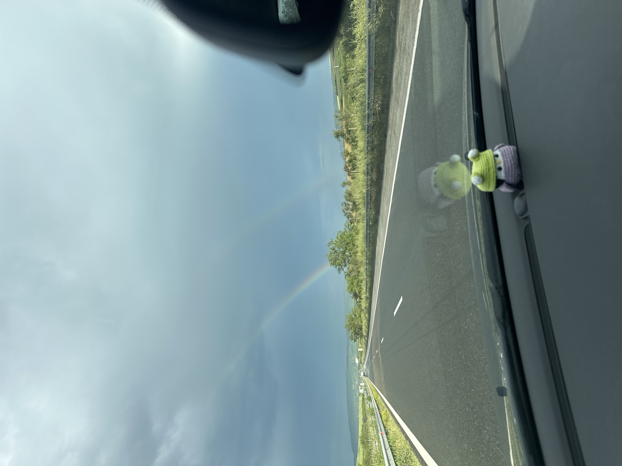 A view from inside a vehicle traveling along a highway. A rainbow is visible in the sky to the left. A small, crocheted owl is attached to the passenger-side window. The location is likely near Mlada Gvardia, Bulgaria. The environment consists of an open highway with greenery on both sides.