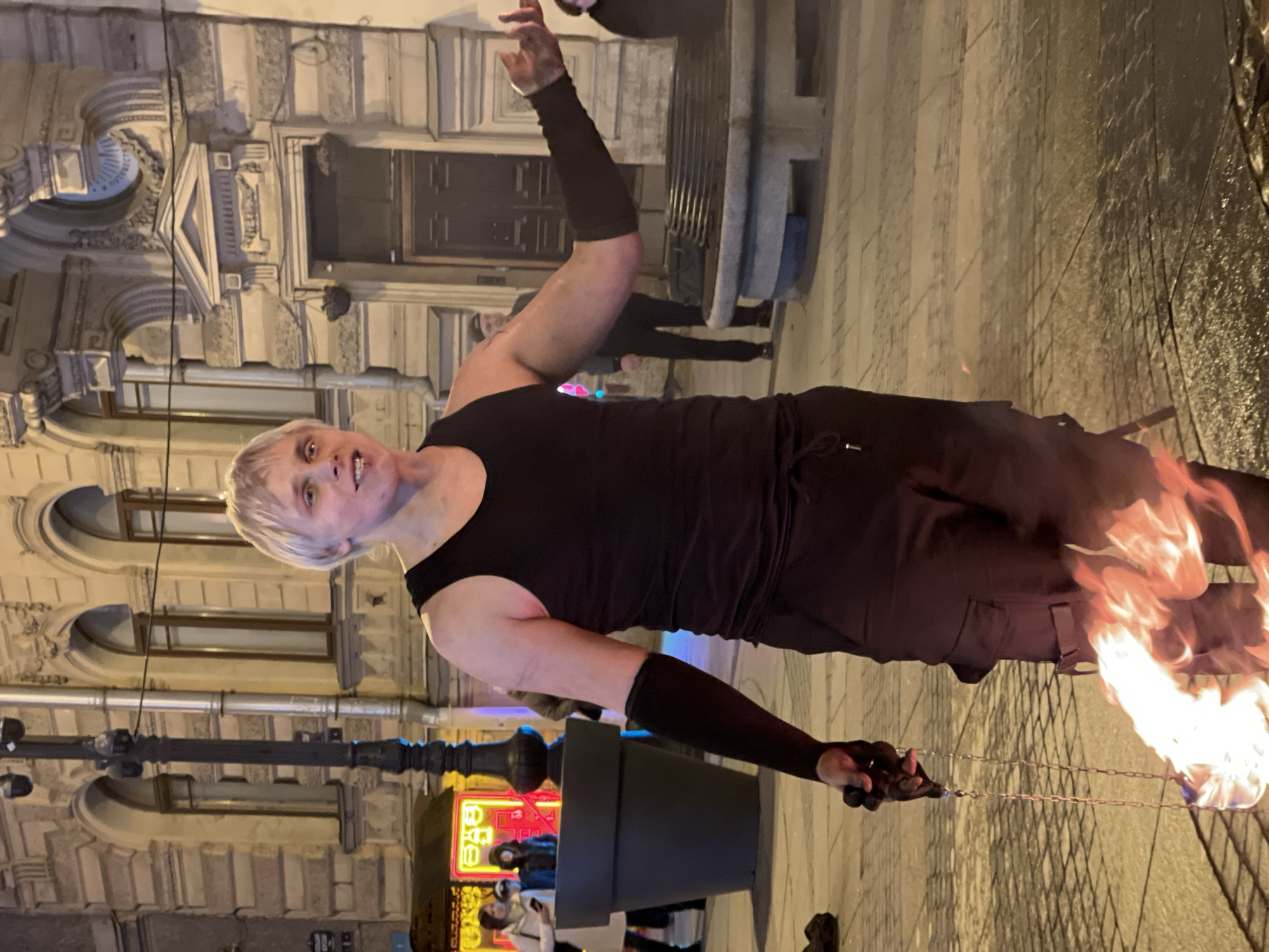 A performer in Saint Petersburg, Russia, is performing with flaming objects. The performer is a fair-skinned individual with short blonde hair, wearing a black tank top, black shorts, and black arm sleeves. They are standing on a cobblestone street, holding chains attached to the flaming objects in their right hand. The flames are bright and dynamic. In the background are ornate building facades and a street lamp with illuminated neon signs. Additional figures can be seen blurred and in the background.