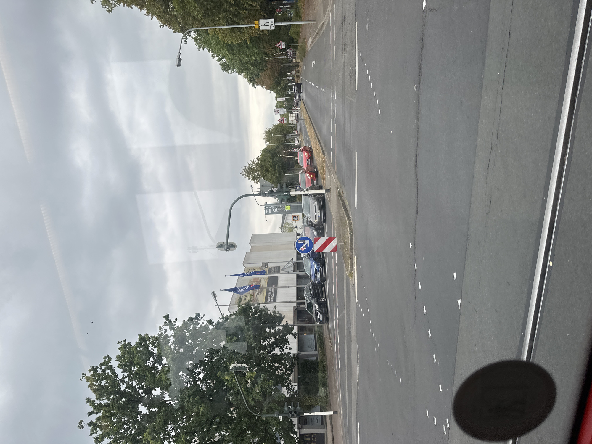 This is a medium-distance, eye-level, forward-facing photograph of a city intersection in Krefeld, Germany, on a cloudy day. The foreground shows a wide asphalt road with white lane markings and reflective road studs. A black, circular object is partially visible in the lower left corner, possibly a reflection or part of the vehicle from which the photo was taken.
In the middle ground, the road continues and curves slightly to the right. Several cars are stopped at the intersection, including a black SUV, a blue sedan, and a red hatchback. A traffic light pole with multiple lights stands on the left side of the road. Across the street, a modern building with large windows and two blue flags is visible. Trees line the right side of the road.
In the background, more trees and buildings can be seen, along with streetlights and traffic signs. The sky is overcast with gray clouds. The image captures a typical urban scene with vehicular traffic and commercial buildings.