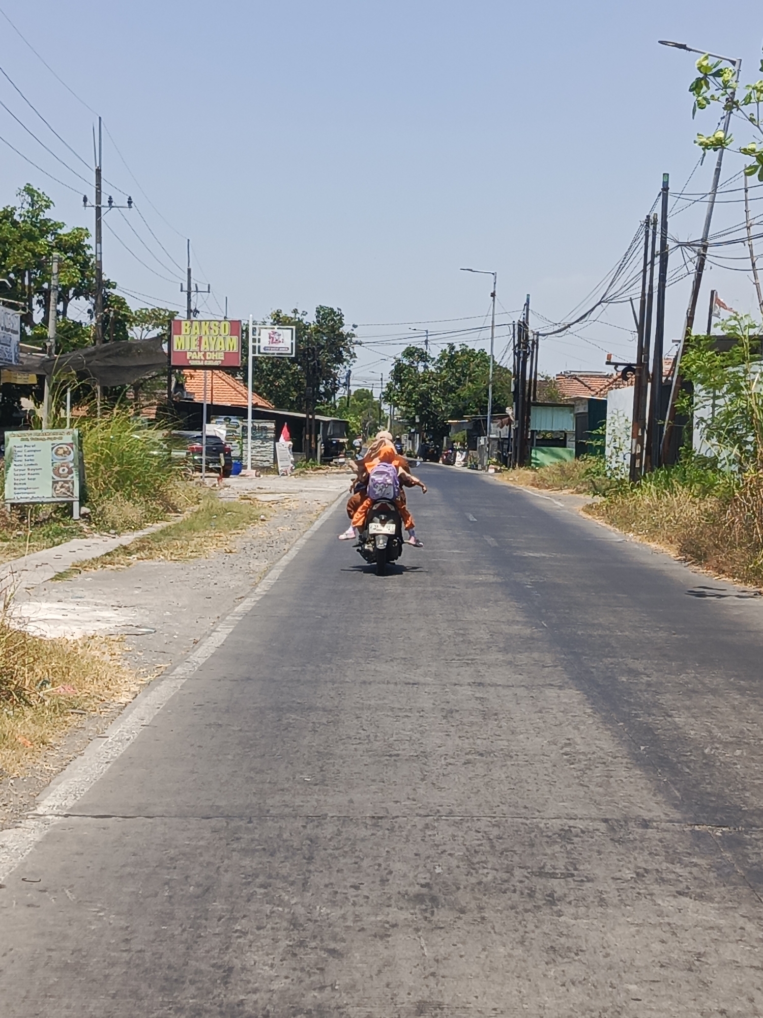 In Prasung, Indonesia, on a clear, sunny day, two individuals are riding on a scooter down a paved road. The person in the front is wearing an orange outfit, while the person in the back is also in orange and has a purple backpack. They are traveling away from the camera. The road is lined with vegetation, buildings, and utility poles. A sign in the distance reads "BAKSO MIE AYAM PAK DHE". A menu board is visible on the left side of the road with text and images of food items. The overall scene depicts a typical day on a street in a Indonesian town.