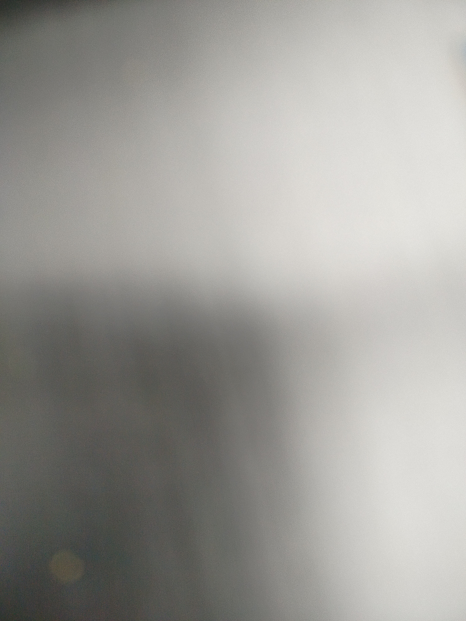 The image is a blurry, close-up shot of a surface, likely indoors. The upper right area is lighter in tone, fading into a darker grey on the left side and at the bottom. There is a pale, out-of-focus circular light reflection in the lower left corner. The texture of the surface appears granular or subtly speckled. There are no discernible people, subjects, objects, actions, or interactions. The environment is undiscernible. The location in Yerevan, Armenia, cannot be determined from the image alone.
