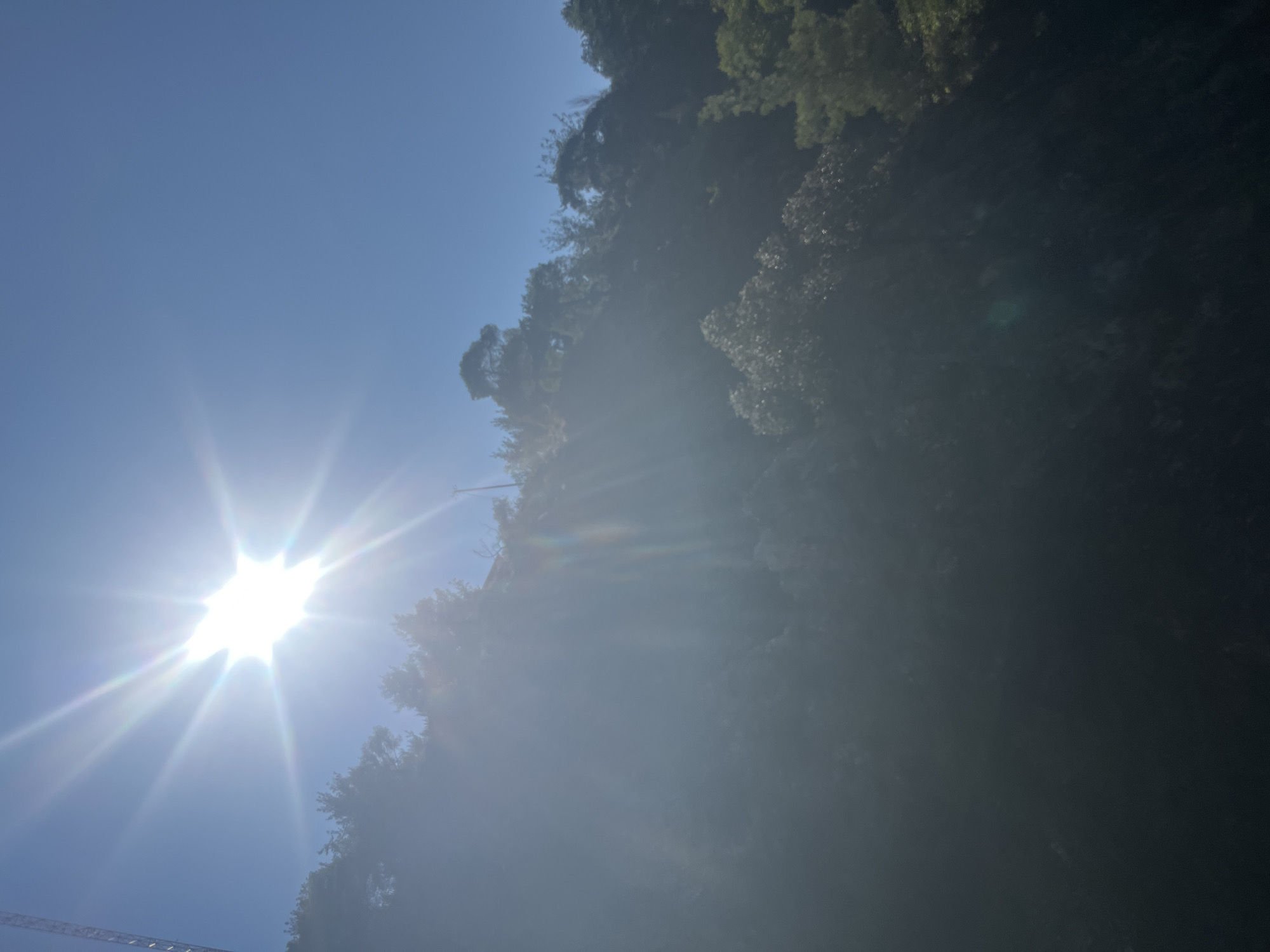 The image captures a bright, sun-drenched sky above a forested hillside in Pano Platres, Cyprus. The sun is the dominant element in the upper left quadrant, emitting strong rays and lens flare that partially obscure the view of the landscape. A portion of a tall, metallic structure, possibly a tower or crane, is visible on the far left edge of the frame. The hillside is densely covered with a variety of trees, their foliage appearing dark and somewhat shadowed due to the intense sunlight. Hints of a building or structures can be discerned nestled among the trees on the upper part of the slope. The overall scene depicts a natural, outdoor environment under clear, sunny weather conditions.