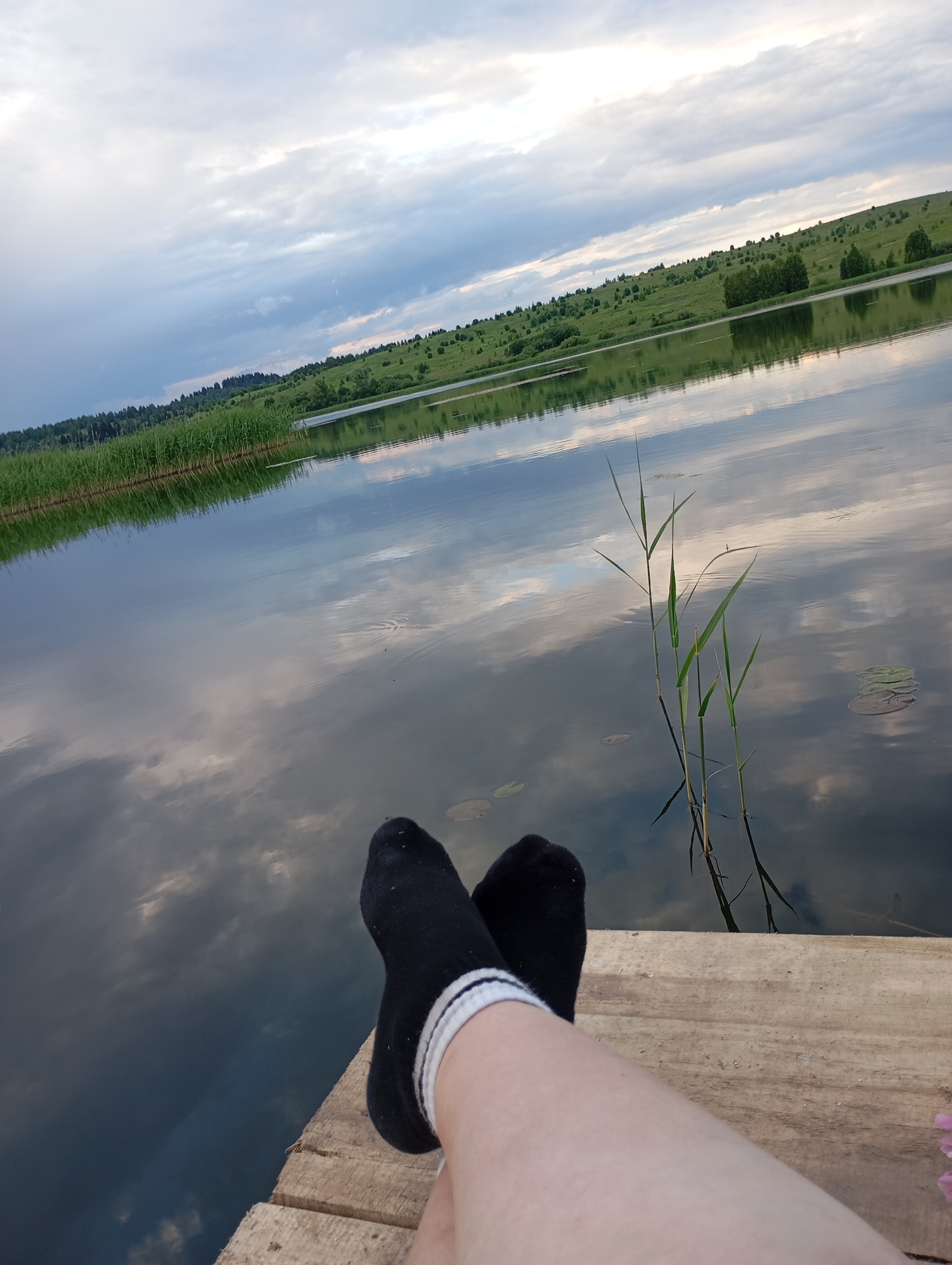 A first-person perspective shot shows a person's legs and feet wearing black socks with white stripes, resting on a wooden dock. The dock extends into a calm lake that reflects the sky. The background features a grassy, rolling hillside dotted with trees and vegetation. The sky is partly cloudy, with soft light indicating either early morning or late afternoon. The overall mood is serene and peaceful, suggesting a moment of relaxation in a natural setting. There is no visible text in the image. The location context provided is Ryabki, Russia.