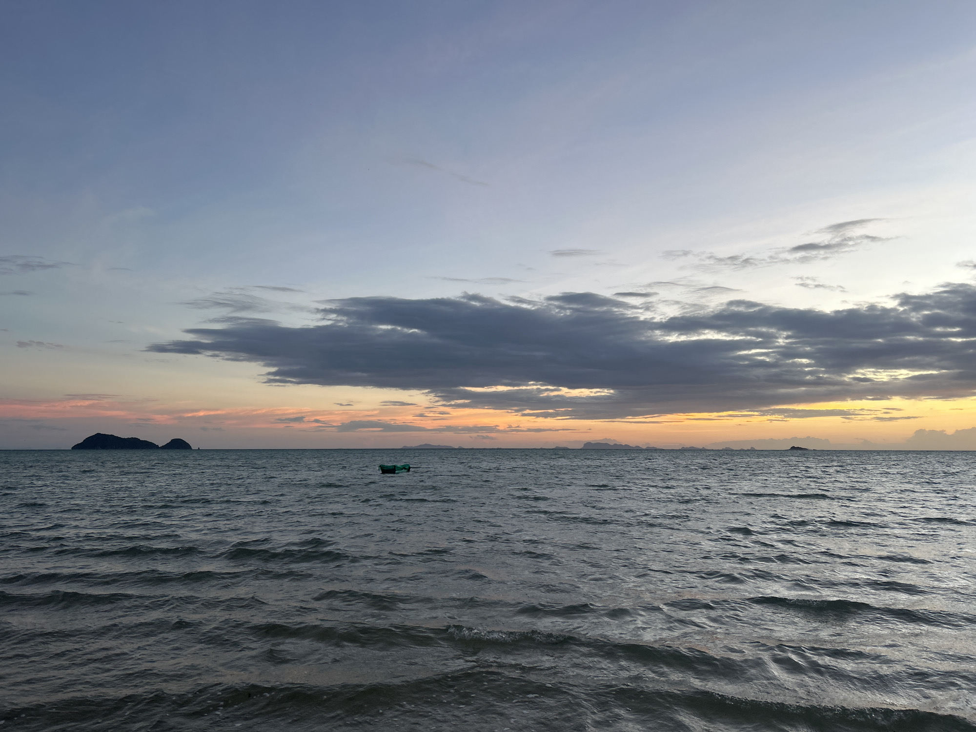The image depicts a serene seascape at dusk in the Ko Pha-ngan District of Thailand. The sky is a gradient of soft blues and grays, transitioning to hues of orange and yellow near the horizon, indicating the setting sun. Dark clouds are scattered across the sky, with lighter streaks of white also visible. The sea is calm, with gentle waves rippling across its surface. In the distance, several islands are silhouetted against the colorful sky, adding depth to the scene. A small, green object, possibly a buoy or a marker, floats in the middle of the water. The overall impression is one of tranquility and natural beauty.