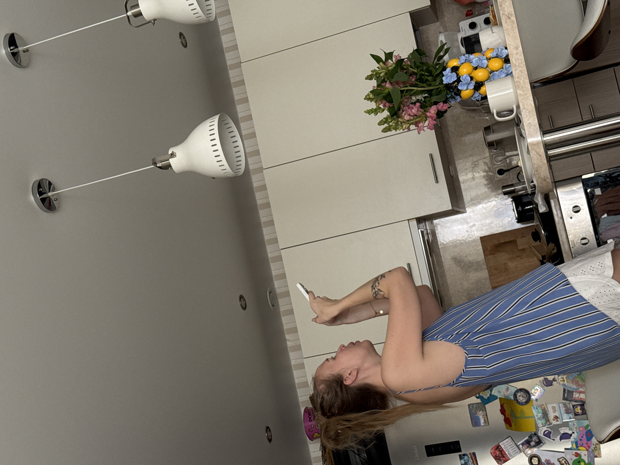 A woman is lying down and looking at her phone in a kitchen setting. She is wearing a blue and white striped dress. The kitchen features white cabinets and a countertop with flowers, fruit, and a mug on it. There are light fixtures on the ceiling, and the refrigerator is covered in magnets. The scene appears to be indoors, likely in a residential setting, but there is no information to confirm that it's set in the city of Sirius, Russia.