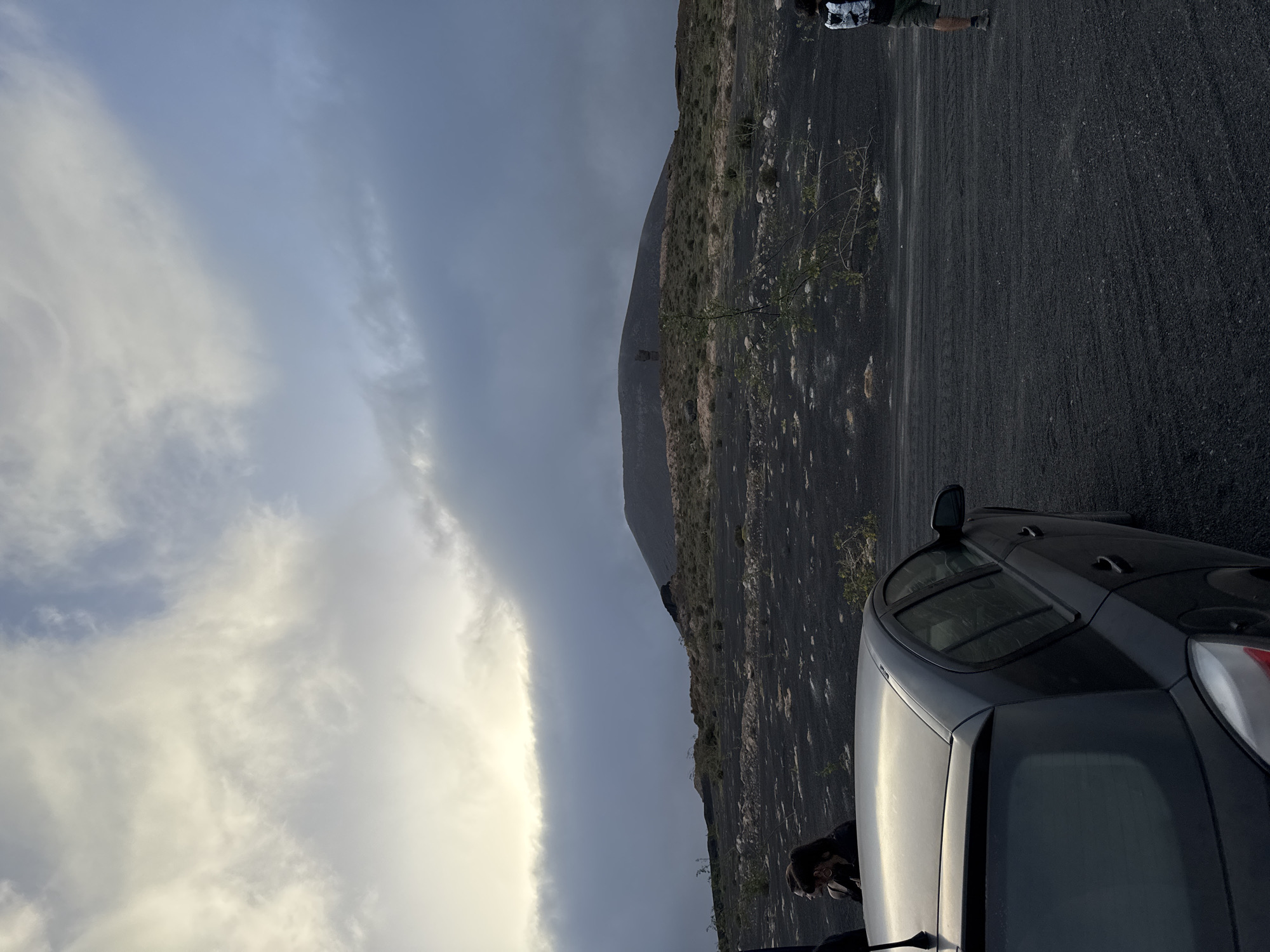 The image depicts a partly cloudy sky over a landscape in Teguise, Spain. In the foreground, a dark-colored car is parked on an asphalt surface. Parts of a person are visible leaning out of the car window. Behind the car, the terrain is rough and volcanic, with sparse vegetation. A mountain or hill dominates the skyline. At the top of the frame, another person is standing on the edge of the terrain. The lighting suggests it's either late afternoon or early morning.