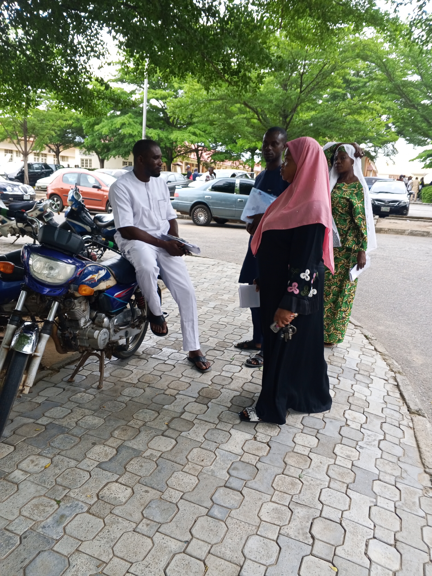 In Lokoja, Nigeria, under a canopy of mature trees, four individuals stand on a paved walkway adjacent to a road. A man, dressed in white traditional attire, is seated on a blue and silver motorcycle. He holds a document in his hands and looks to his right. Beside him, to his left, a woman in a pink hijab and a long black dress, adorned with floral embroidery on the sleeves, holds a set of keys. To the right of the woman in pink, another woman, wearing a green and gold patterned dress and a white headscarf, stands with a document in her hand. A fourth individual, a man in a dark shirt and light trousers, stands slightly behind the woman in the pink hijab, also holding a document. In the background, several cars are parked or driving on the road, including an orange sedan, a silver sedan, and a black SUV. The environment is a public outdoor space, likely near a building or administrative center, given the presence of people with documents and the parked vehicles. The paved walkway is constructed of interlocking gray pavers.