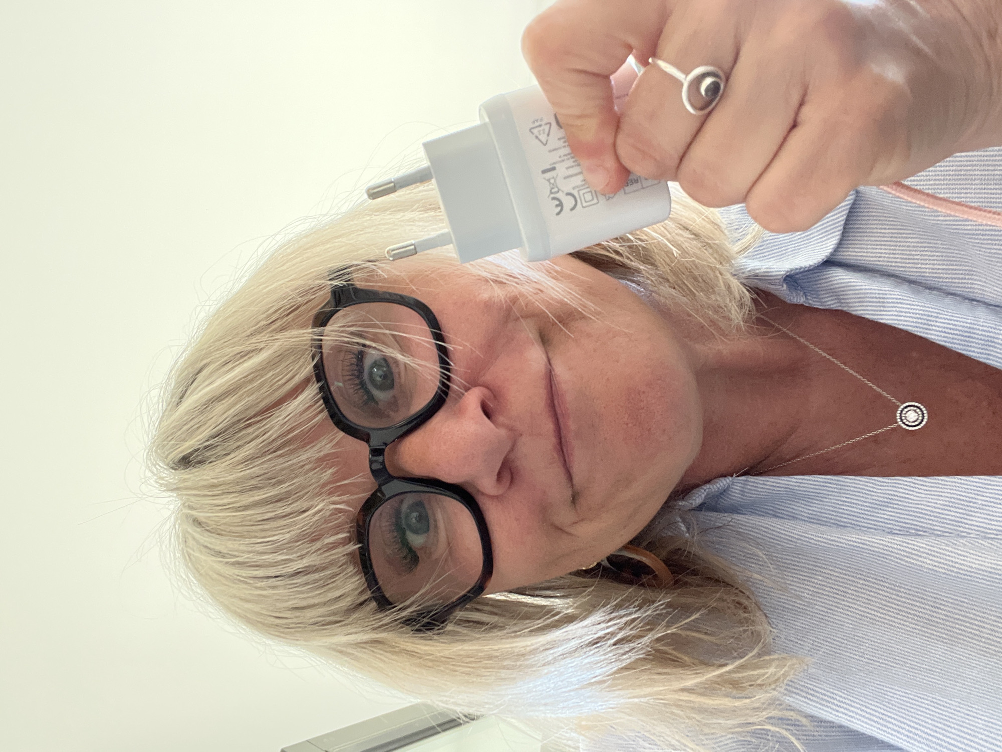 A woman with blond hair and glasses holds a white European-style electrical plug. She is wearing a light blue striped shirt and a silver necklace with a circular pendant. A silver ring with a circular design is on her right hand. The image does not contain enough detail to determine the location as Chambœuf, France or provide a more detailed forensic description.
