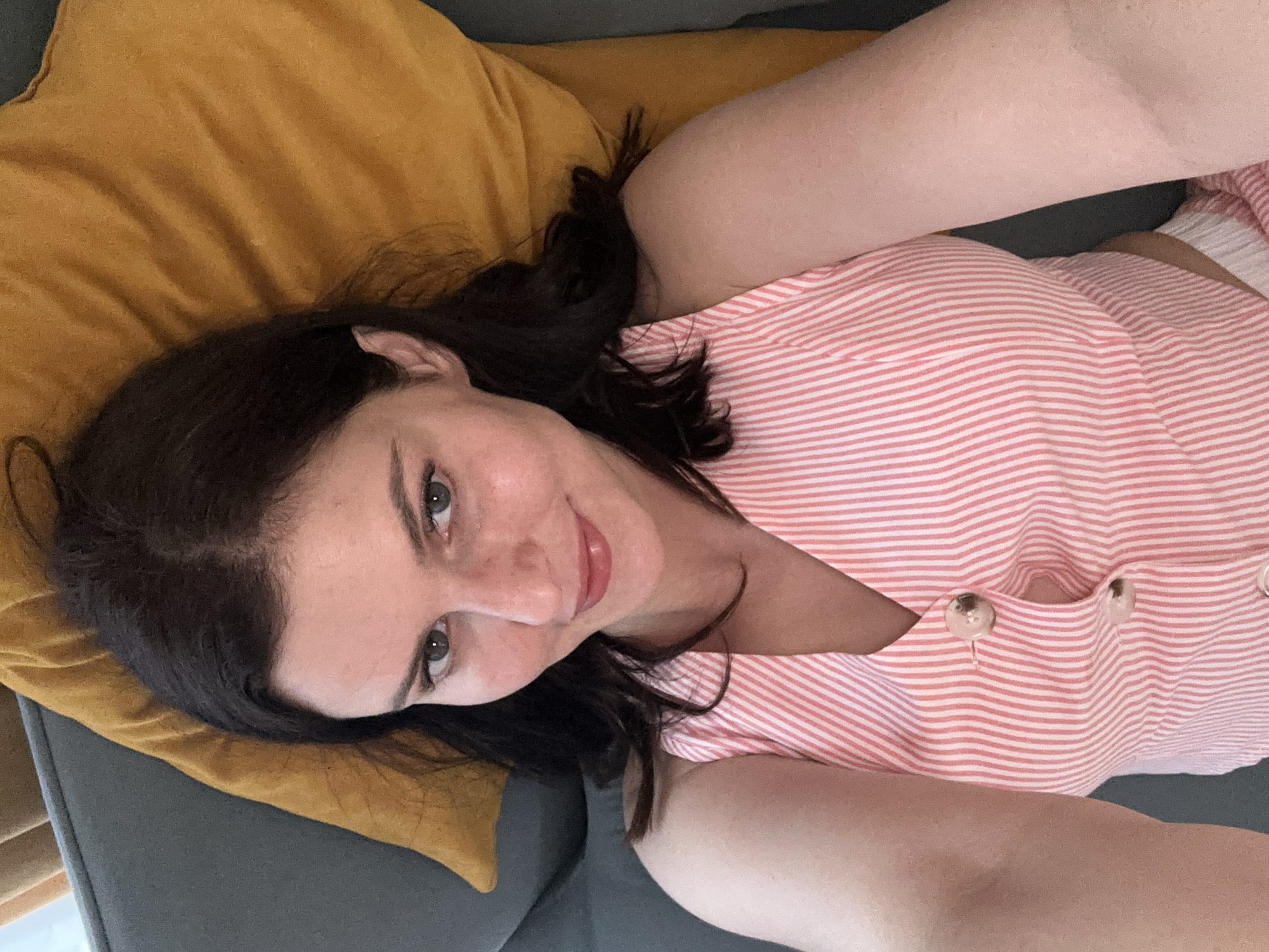 A woman with dark hair and light skin is lying down, looking at the camera. She is wearing a pink and white striped top. She has light green eyes and is smiling. She is lying on a couch with a yellow pillow behind her head. The lighting suggests it is daytime.