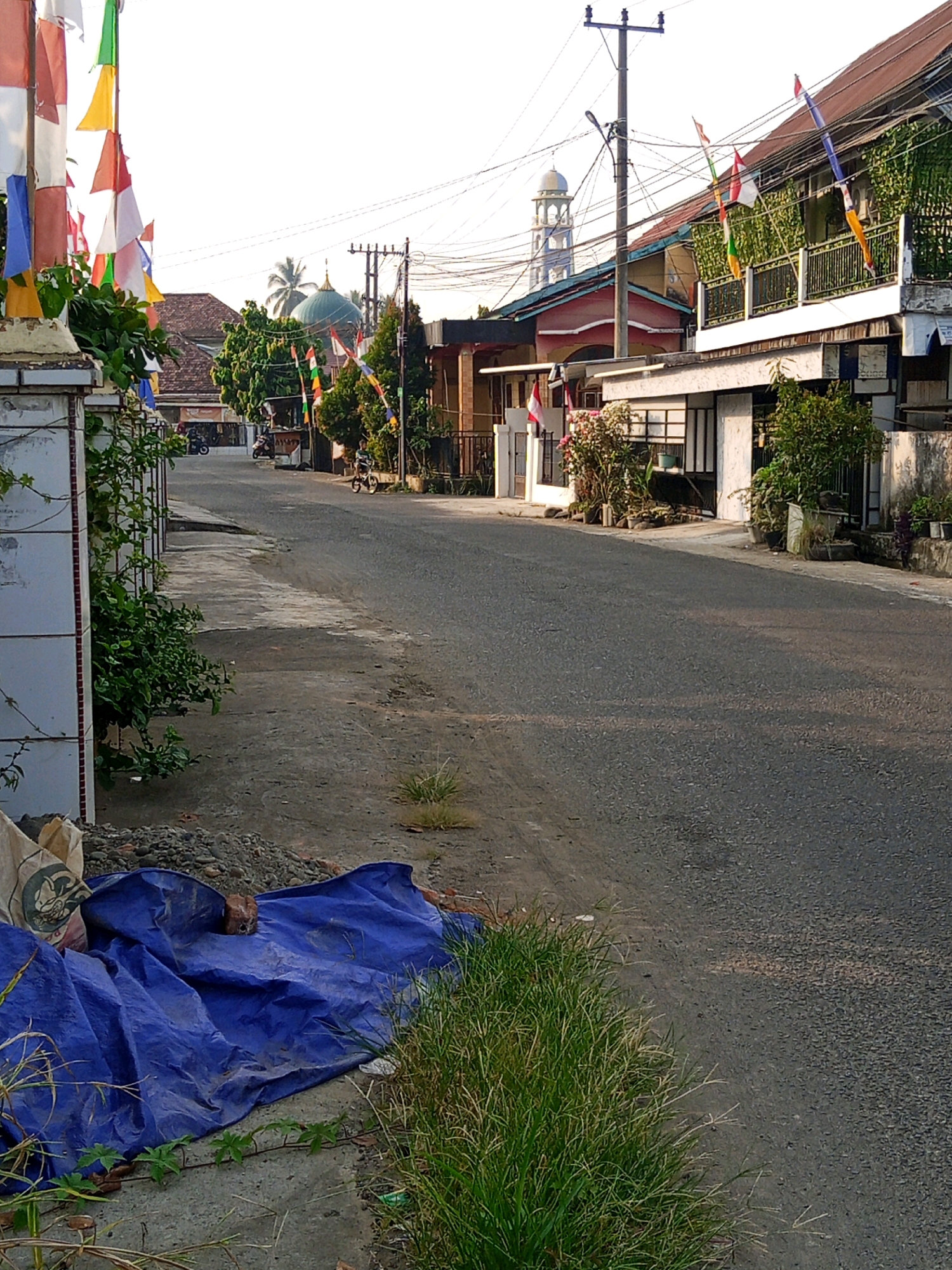 The image depicts a street scene in the city of Lubuklinggau, Indonesia, likely during daylight hours due to the bright sky and the shadows cast. The street itself is paved and appears to be a two-lane road. On the left side of the road, there is a white tiled wall with green foliage growing on it and some small flags attached. Further down the street, there are houses with red-tiled roofs and balconies, some adorned with ivy. A mosque with a green dome and a minaret is visible in the distance, suggesting the location is within a residential area. On the right side of the street, more houses are visible, with balconies and vegetation. A few motorcycles are parked, and a child is riding a bicycle on the street. In the foreground on the left, there is a pile of gravel and a large blue tarp covering something, along with a sack. Green grass grows along the edge of the road. Overhead, power lines and a utility pole are present. The overall impression is a quiet street in a tropical or subtropical urban environment.