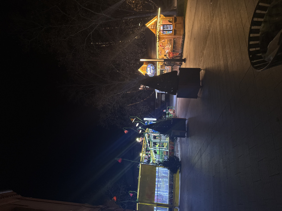 A nighttime scene shows a street fair. A wooden stall with gingerbread details stands near a Christmas tree. Further down, a children's ride is illuminated with lights shaped like flowers. Bare trees are decorated with string lights. The ground is paved, and the sky is dark. A blurry object appears at the top right.