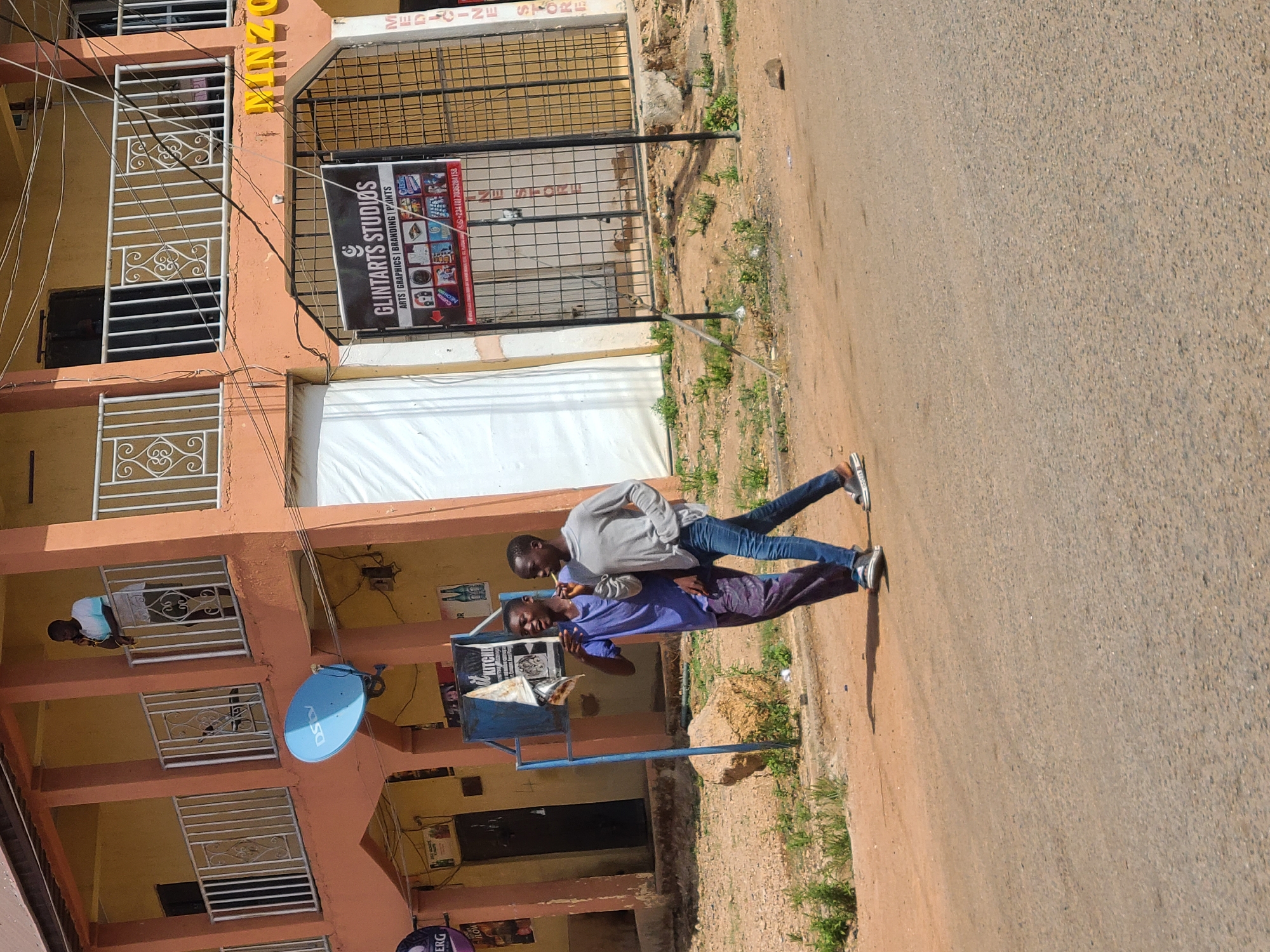 This media file captures a vibrant street scene in Jos, Nigeria, illuminated by bright daylight, likely mid-morning or afternoon given the clear shadows.
The primary subjects are two young men standing on a dirt shoulder beside a paved road. One man, on the left, wears a light grey long-sleeved shirt, blue jeans, and sneakers, looking towards his companion. The second man, on the right, sports a purple short-sleeved shirt, patterned trousers, and sneakers, and appears to be speaking or reacting to the other. Their interaction seems casual and relaxed.
To the left, a multi-story building with a light peach-colored facade dominates the background. It features white metal grilles on its windows and balconies. A blue DStv satellite dish is mounted on the building. On one of the upper balconies, another person in a white top is visible. Numerous power lines crisscross the front of the building.
Visible text on signs attached to the building includes "GLINTARTS STUDIOS," advertising "ARTS / GRAPHICS | BRANDING | PAINTS," along with a contact number: "+234 (0) 7036284158." Another sign, partially visible, indicates a "MEDICINE STORE." Other text snippets like "ERG," "ROW," and "KITCHEN" are also discernable on posters or signs. Near the two men stands a blue metal frame, possibly supporting a waste bin. The ground is a mix of reddish-brown dirt, sparse green vegetation, and grey asphalt on the road.
