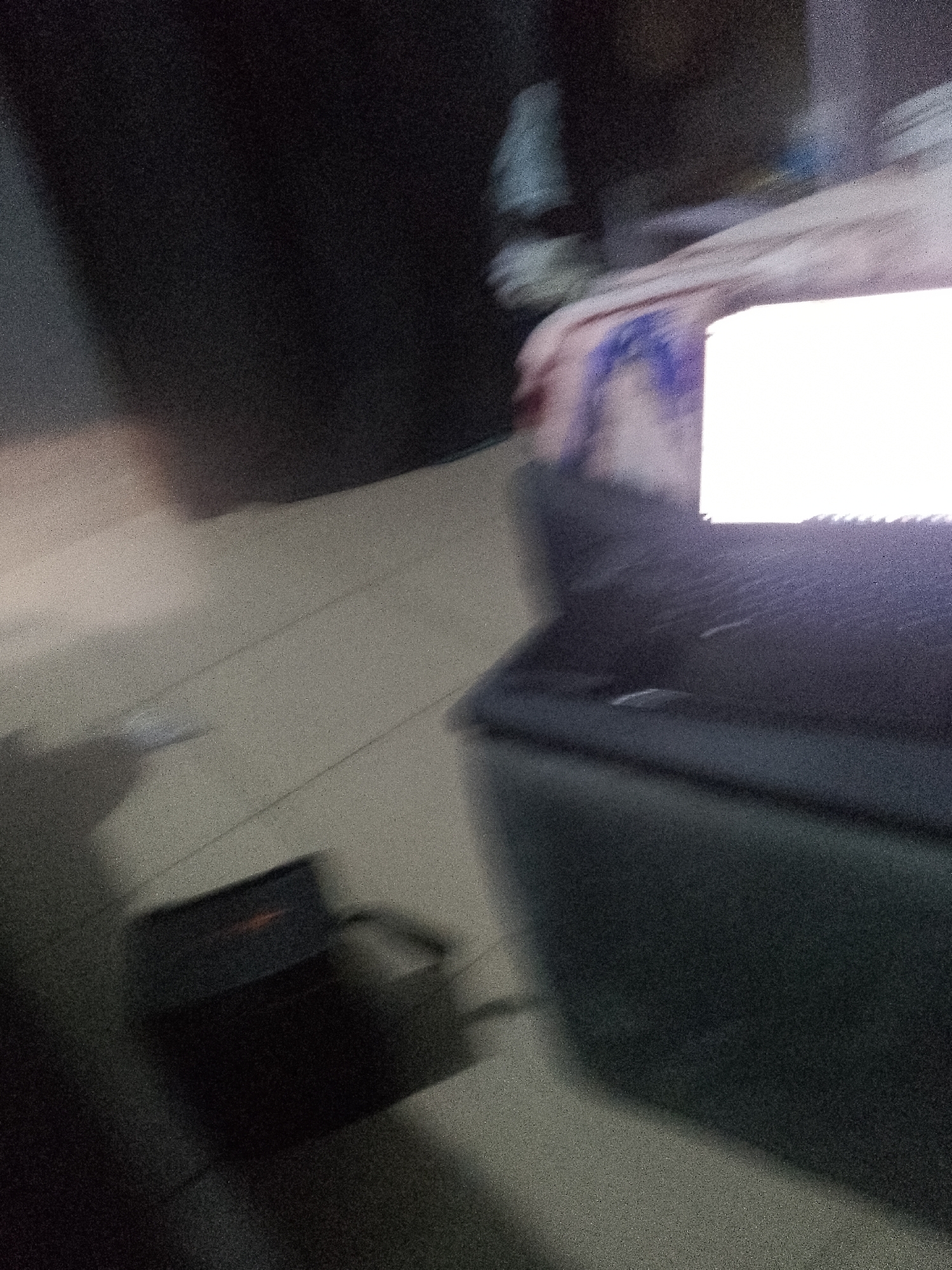 The image is a blurred, low-light, interior shot that appears to be taken with a mobile phone. The primary subjects are a laptop with a bright white screen and a small black electronic device with a red indicator light, possibly a power adapter, situated on a tiled floor. The background is dark and indistinct, with hints of fabric and possibly a person's arm in motion. The overall impression is one of a hastily taken photograph, likely in a residential or office setting. The location in Jauro Yinu, Nigeria, is not visually discernible from the image itself, as there are no identifying landmarks or contextual cues. The image is heavily affected by motion blur, rendering details unclear.